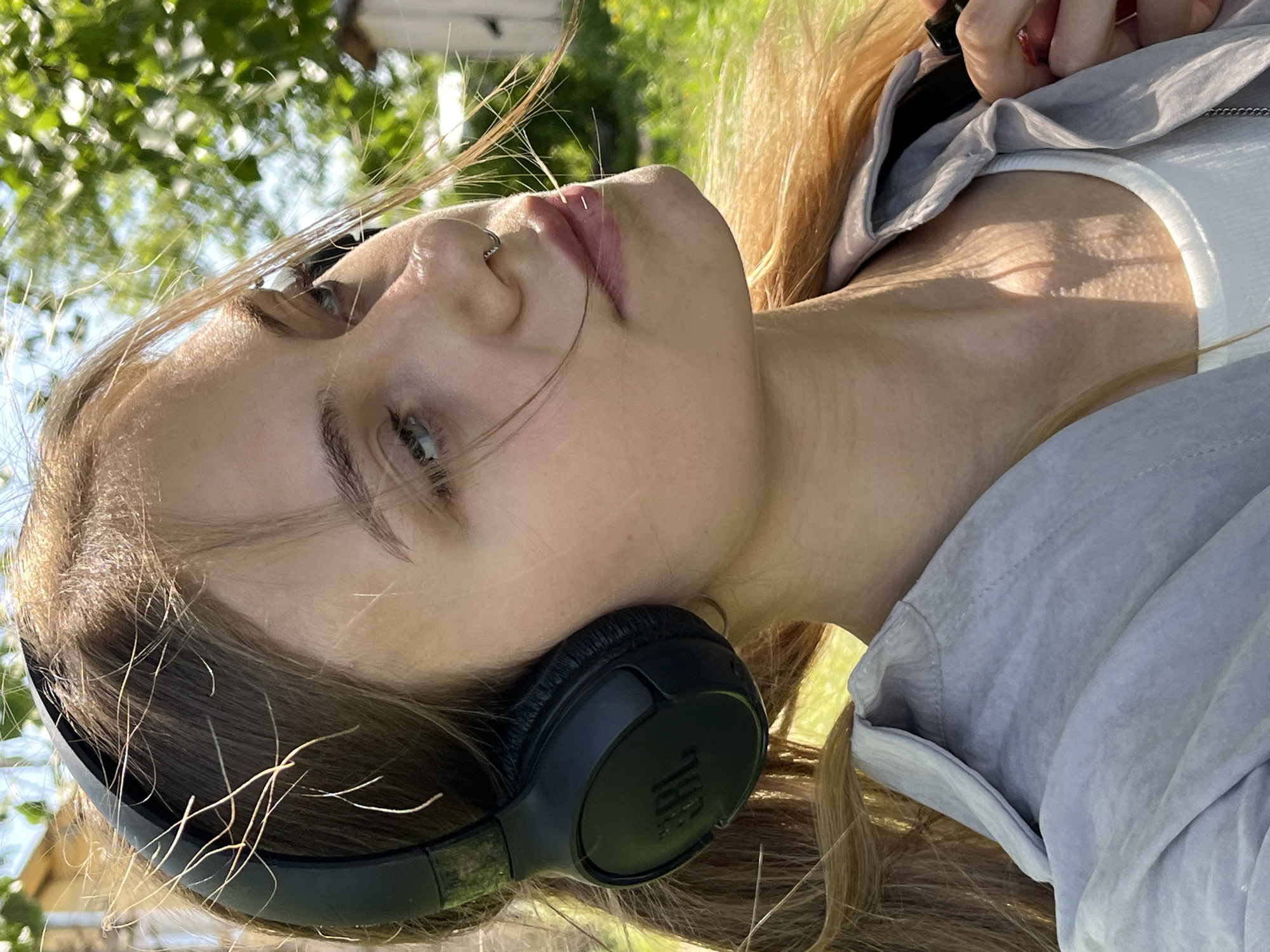 A young woman lies on her back outdoors, wearing black JBL headphones. She has fair skin, light brown hair, and a nose ring. She is wearing a white top and a light gray overshirt. The background is blurry, but appears to be foliage and trees, suggesting a park or outdoor area. The location provided, Фоки, Russia, is not identifiable within the visual data of the image.