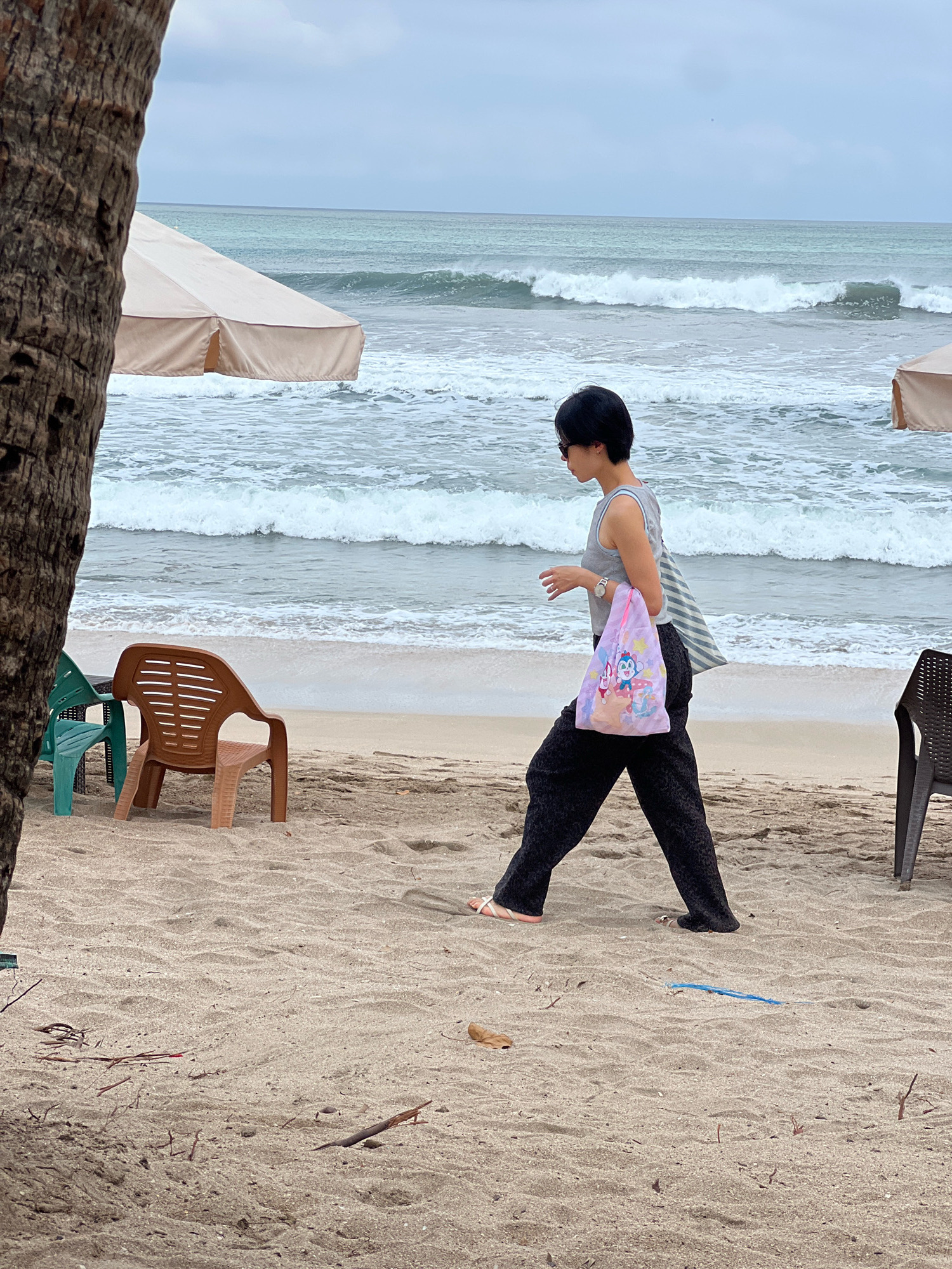 A woman walks on a sandy beach in Kuta, Indonesia, beside the ocean. The woman, wearing sunglasses, a gray tank top, and black pants, carries a light-colored tote bag with a cartoon pattern on her left shoulder. She is wearing white sandals. Behind her, the ocean has moderate waves and a cloudy sky. Two beach umbrellas are visible, one partially obscured by a palm tree trunk on the left, and another on the right. Several plastic chairs are scattered on the sand.