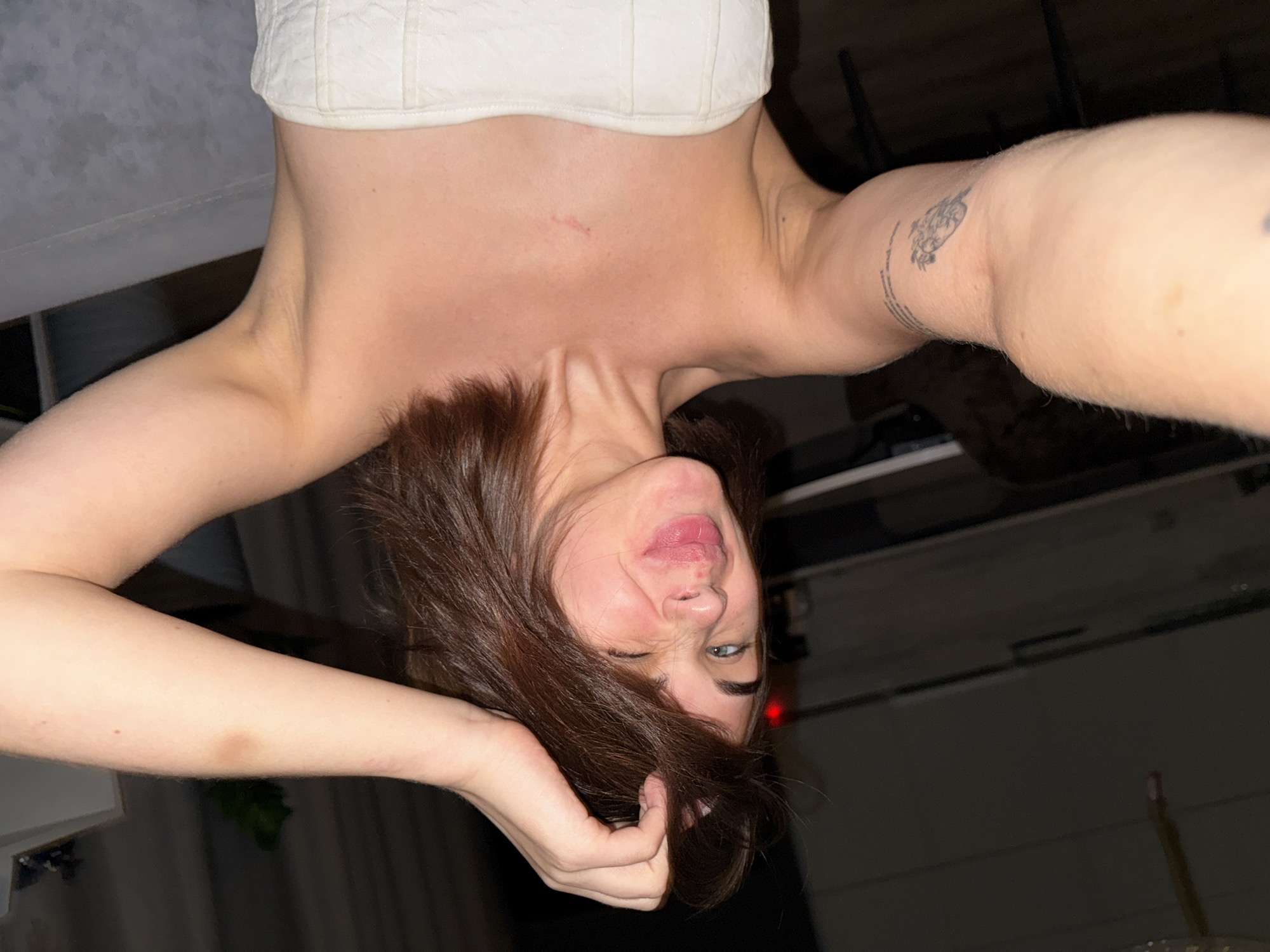 A person with medium-length brown hair is positioned upside down, taking a selfie indoors. The person is wearing a white top. The background is dimly lit, and some objects are visible, including what appears to be furniture. The location appears to be indoors and cannot be verified to be Лобаново, Russia.