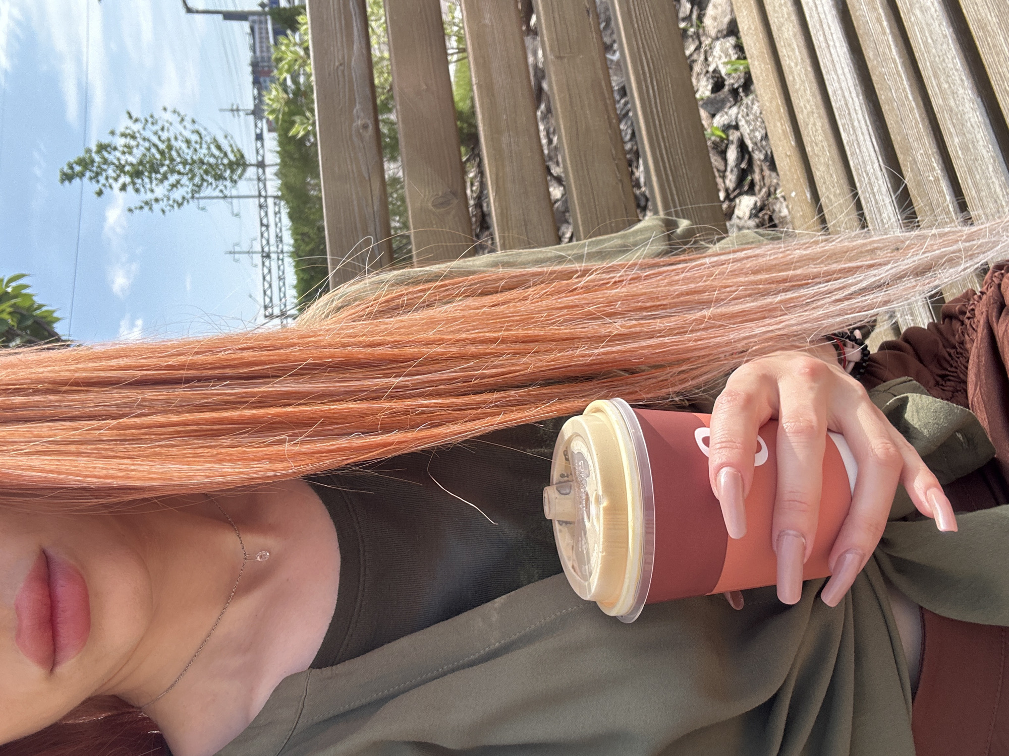 A medium shot captures a person's upper body as they sit on a wooden bench outdoors. The person has long, dyed ginger hair that cascades down their chest. They are holding a red and tan disposable coffee cup in their right hand. Their fingernails are long and painted a pale pink. The person is wearing a green long-sleeved shirt over a dark green t-shirt. The background shows a fence, trees, and electrical poles against a bright blue sky with scattered clouds. The lighting suggests it is daytime. The location is identified as Unknown, Unknown.