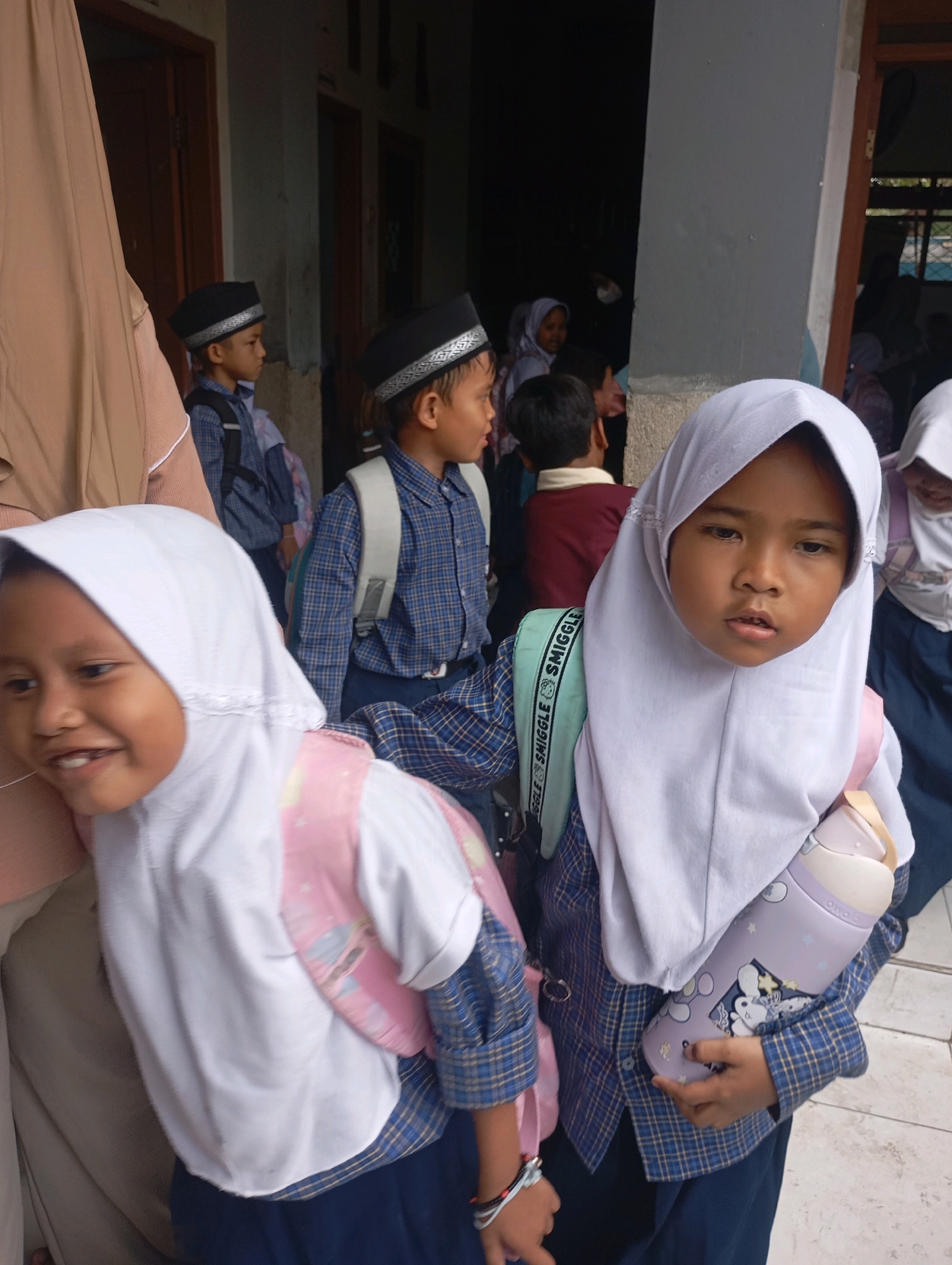 The image depicts a group of children, likely students, outside a building in Babelan, Indonesia. Two girls in the foreground are wearing white hijabs. The girl on the left is smiling broadly, while the girl on the right looks directly at the camera with a neutral expression. Both girls are wearing blue plaid shirts and dark blue skirts, and have backpacks. The girl on the right is holding a light purple water bottle decorated with cartoon characters. Behind them, other children are visible, some wearing similar attire and backpacks, and some wearing black caps. The background shows a building entrance with wooden doors and a gray wall. The environment suggests an outdoor school setting.