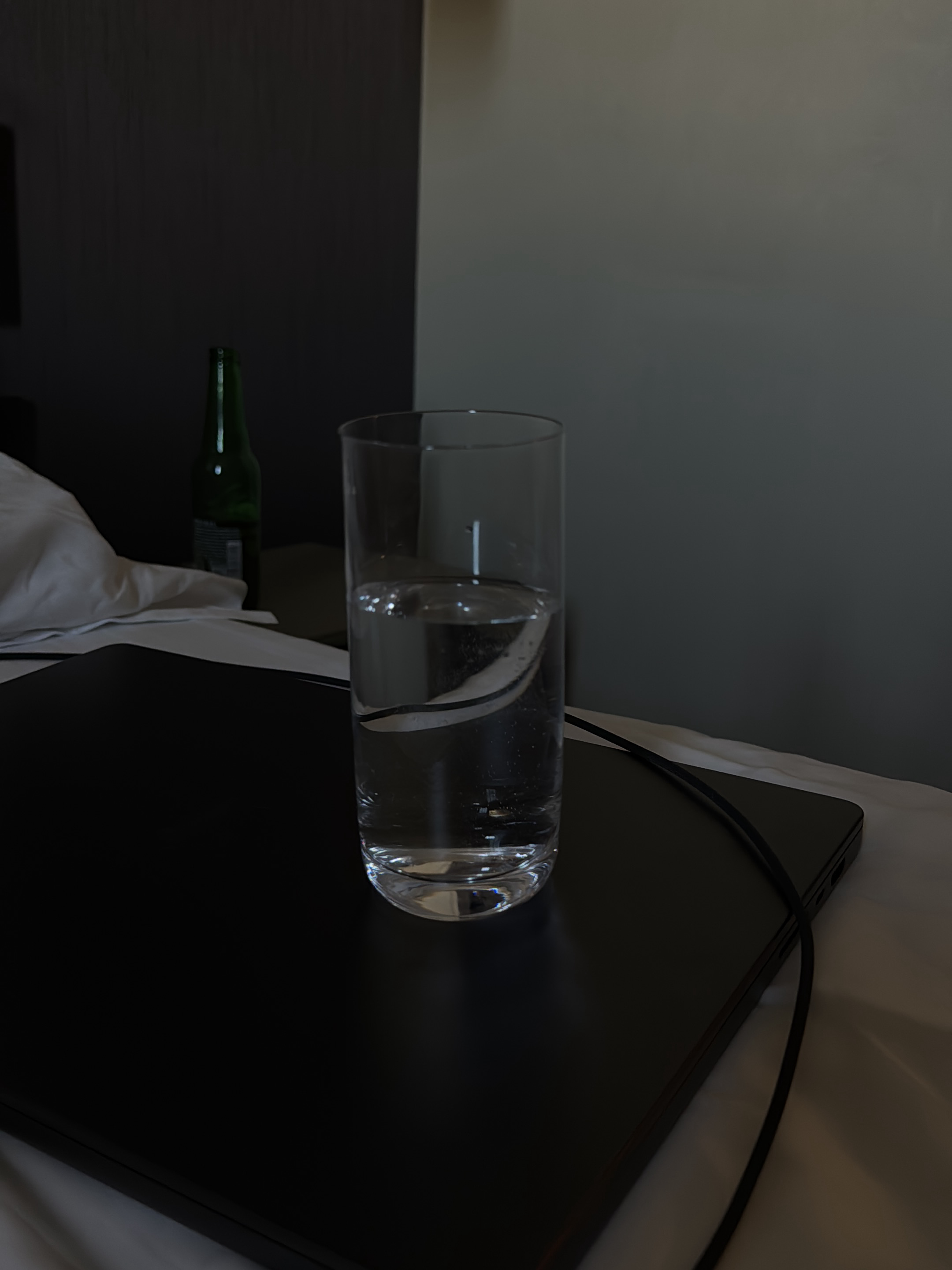 An indoor scene, likely a hotel room in Milan, Italy, features a clear glass of water filled about two-thirds full, placed on a closed black laptop. The laptop rests on white bedding. To the left of the glass, a dark green bottle, likely a beer bottle, stands upright. The background consists of a dark, textured wall on the left and a lighter, smooth wall on the right. A black cable is visible, draped across the laptop and extending out of frame. The lighting is dim, casting shadows and highlighting the water's movement within the glass.