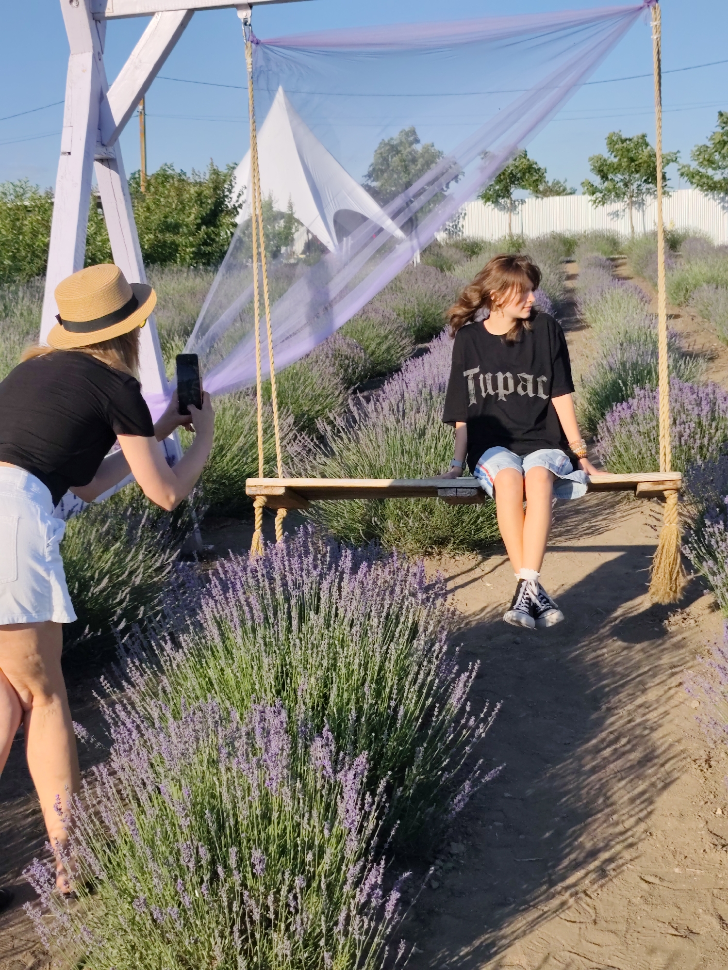 A young woman with brown hair is sitting on a wooden swing in a lavender field. She is wearing a black t-shirt with "Tupac" written on it in white letters, and checkered shorts. She has on black high-top sneakers. Her right arm is resting on the swing and her left arm is bent at the elbow. Another woman, wearing a straw hat and a black t-shirt and white shorts, is standing to the left of the swing and taking a photo of the seated woman with her phone. The swing is suspended from a light purple wooden A-frame structure, draped with sheer lavender fabric and a white triangular cloth. The lavender field stretches out behind the swing. The ground is dirt. The sky is clear and blue, and the sun is casting shadows from the lavender plants and the swing structure. The scene appears to be outdoors, likely in Nechaiane, Ukraine, given the context.