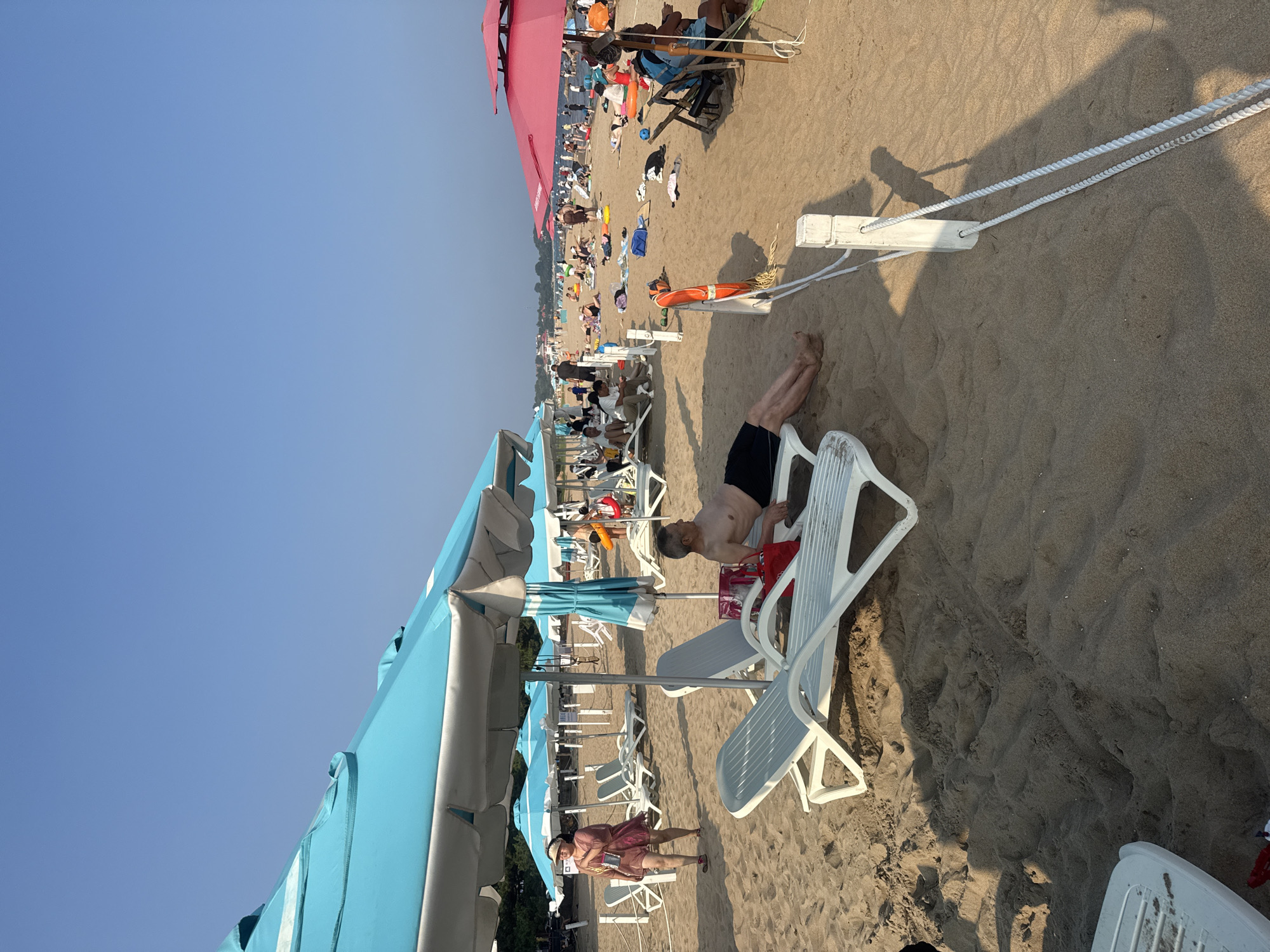 The image depicts a crowded beach scene in Qinhuangdao, China, likely during daytime. Several beach umbrellas, predominantly blue and pink, are set up on the sand. Numerous people are visible, some sunbathing on lounge chairs, while others are walking or scattered across the beach. In the foreground, a man is relaxing on a white lounge chair, wearing black shorts. To the left, a woman in a pink dress is walking, carrying a bag. In the mid-ground, more lounge chairs and umbrellas are arranged, with people seated or lying down. Further in the background, a larger gathering of people is visible near the water, suggesting swimming or other beach activities. The sand appears light brown, and the sky is clear and blue. The overall impression is a lively and busy beach environment.
