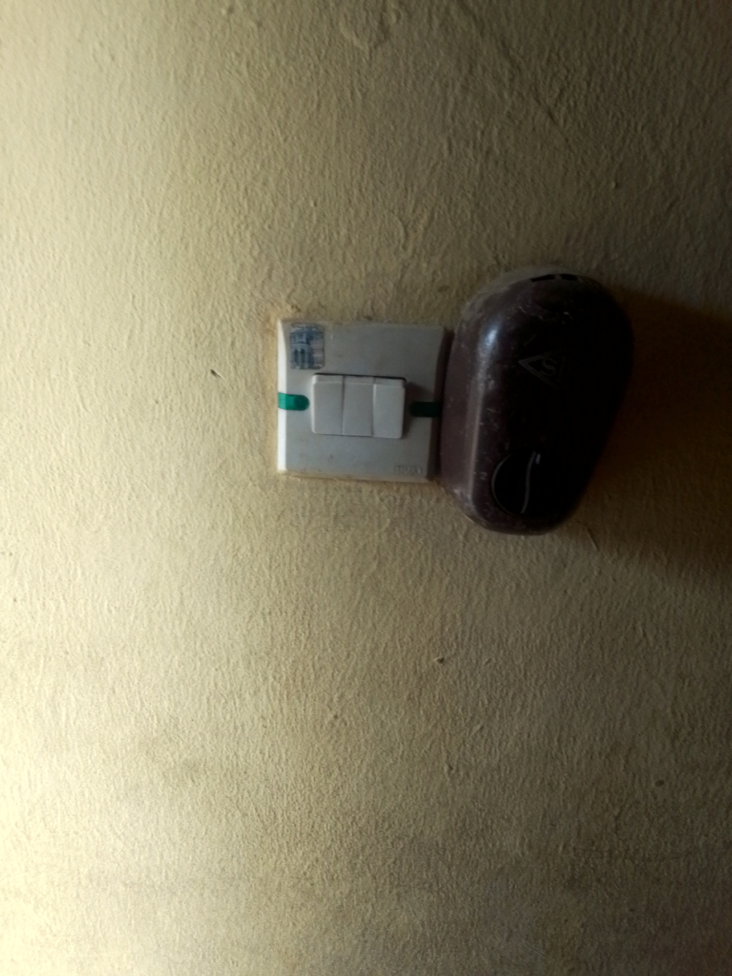 The image shows a close-up of a light switch and a dark, bulbous object attached to it, mounted on a textured, pale yellow wall. The light switch has three white toggles, with the middle one appearing to be in the "on" position. A small green piece of plastic is visible, possibly a label or indicator, to the left of the toggles.
The dark object, which obscures a portion of the light switch, is smooth and appears to be made of plastic. It has a distinctive shape, wider at the top and tapering towards the bottom, with a curved outline. A small symbol, resembling a triangle with an "S" inside, is visible on its surface, along with some faint numbering or markings. The object is angled towards the viewer, casting a shadow on the wall.
The wall itself has a rough, uneven texture, typical of a plastered surface. The lighting in the scene is dim, with a strong light source coming from the left, creating shadows and highlighting the texture of the wall. The overall impression is of an indoor setting, possibly a home or a public building, in Jalingo, Nigeria. There are no people visible, and the activity is static, focusing on the electrical fixture. The visible text consists of the symbol and faint markings on the dark object.