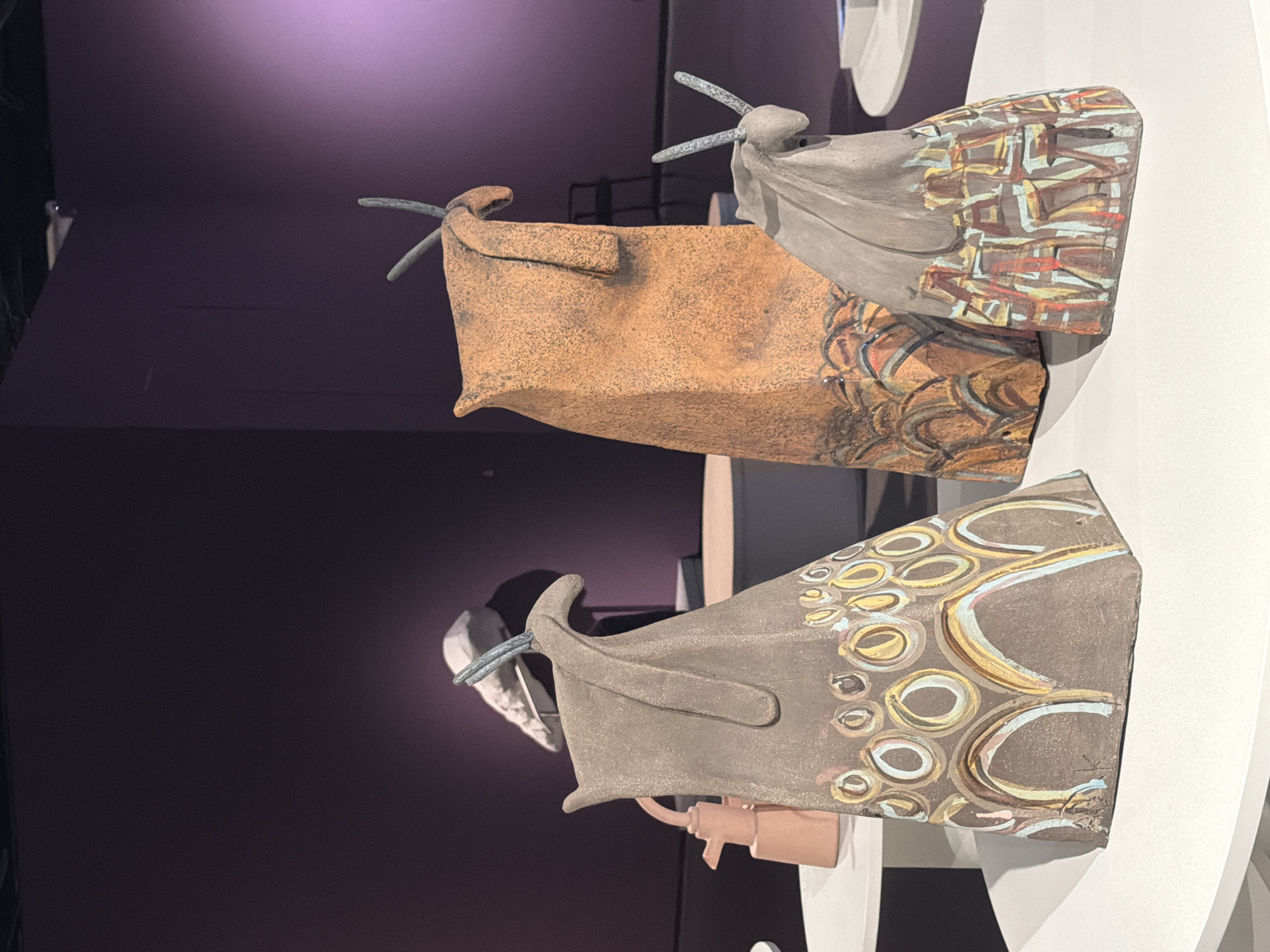 The image shows three ceramic sculptures on display in a modern art museum. The sculptures are abstract, resembling stylized animal heads or boats with rope-like antennae. The sculptures are painted with patterns: one has a grey body and a pattern of multicolored circles, another is brown with a pattern of swirled lines, and the third is grey with a linear geometric pattern. The sculptures are displayed on a white, circular pedestal. The background is a dark purple wall. The location is likely an art museum in Tel-Aviv, Israel, given the style of art and context.
