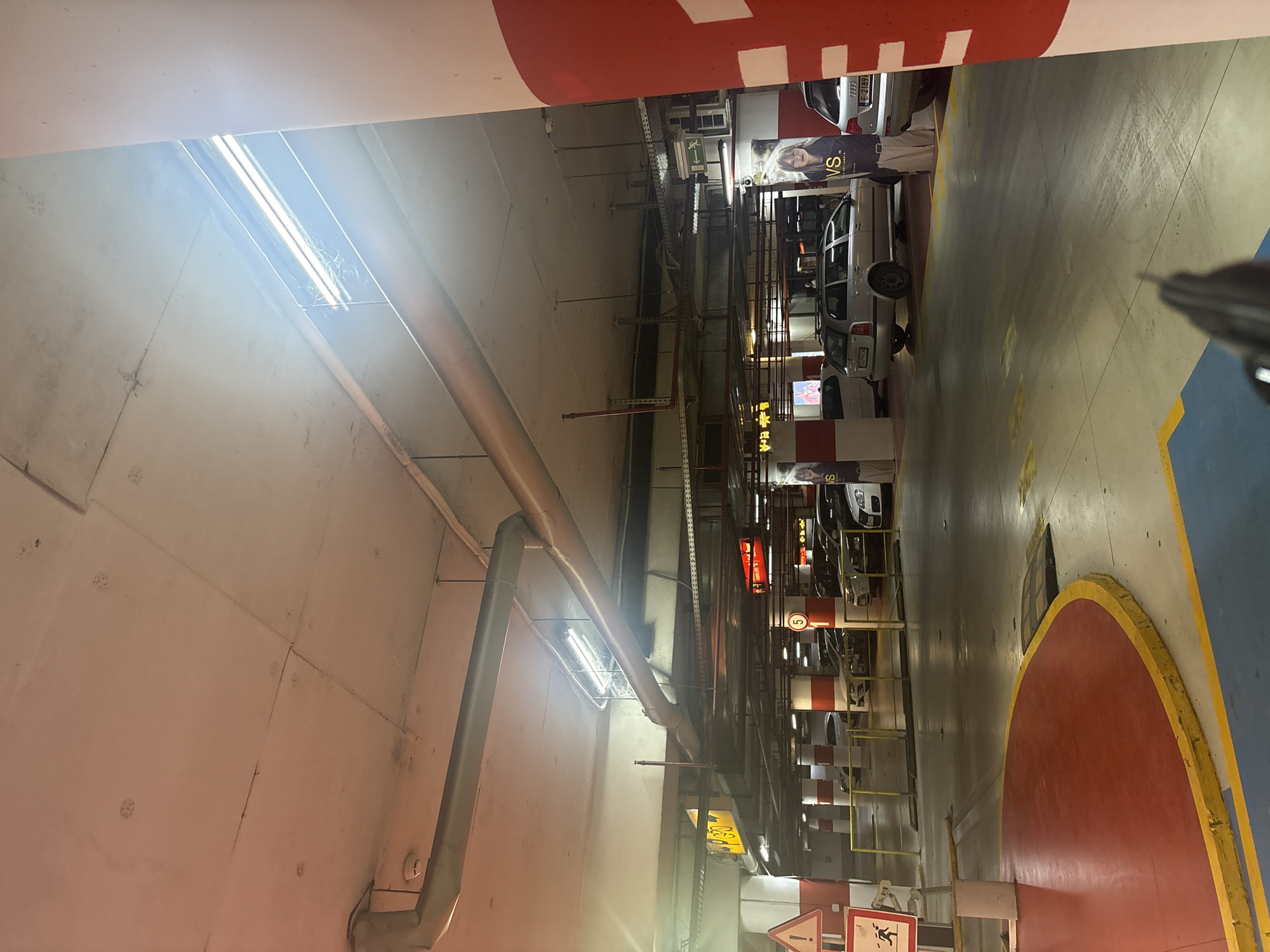 The image shows an interior view of a multi-level parking garage. The ceiling is made of concrete, with exposed pipes and fluorescent lighting fixtures. Several cars are parked in designated spots. Pillars painted with red and white stripes support the ceiling. In the foreground, there are colored markings on the floor, including a red circular area. Signs are visible, including one with a running figure and a triangular warning symbol, and another circular sign with the number "5" inside a red circle. Posters are displayed on some of the pillars, featuring images of models with the "VS" logo. The lighting suggests it is daytime, with sunlight filtering in from a higher level.