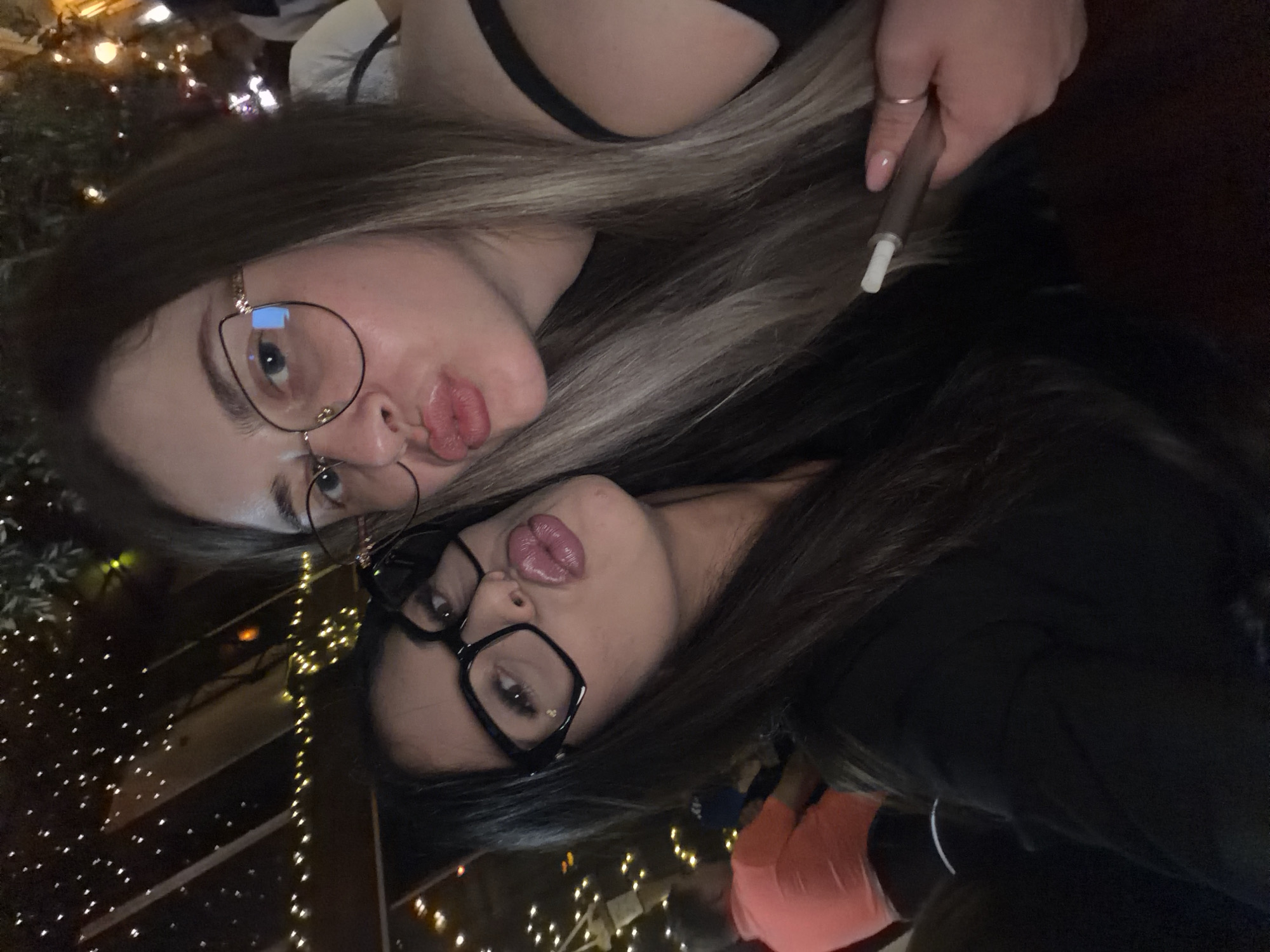 Two young women are taking a selfie in what appears to be an outdoor or semi-outdoor venue, possibly a restaurant or bar, at night. The background shows decorative lighting strings and foliage. The woman on the left has long dark hair and is wearing black-rimmed glasses. She is leaning in towards the camera, with her lips puckered. The woman on the right has lighter, blonde-streaked hair and is also wearing glasses. She is also leaning in, with her lips puckered, and has a slender object, possibly an e-cigarette or vape pen, in her right hand. In the blurred background to the left, a man in an orange shirt is visible, suggesting other patrons are present. The overall ambiance is casual and social. There is no specific information in the image to identify the location as Plovdiv, Bulgaria.