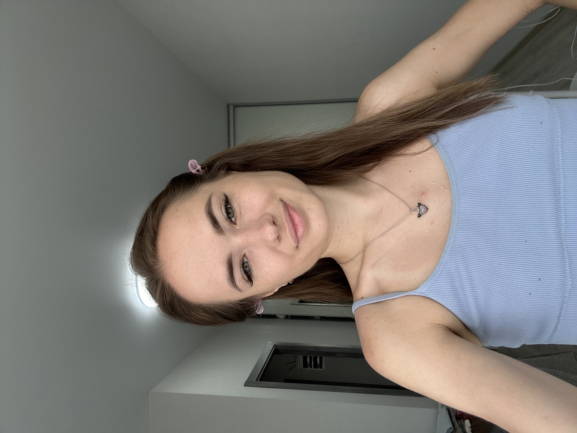 A young woman with long brown hair is pictured indoors, facing forward and looking towards the camera. She is wearing a light blue ribbed tank top and a silver necklace with a small heart-shaped pendant. Her hair is partially clipped back with pink hair clips on either side of her head. She has light skin with freckles, and is wearing light makeup, including mascara and eyeliner. Her expression is a gentle smile. The background appears to be a hallway with white walls, a light fixture on the ceiling, and doors visible on either side. The overall lighting is soft and natural.