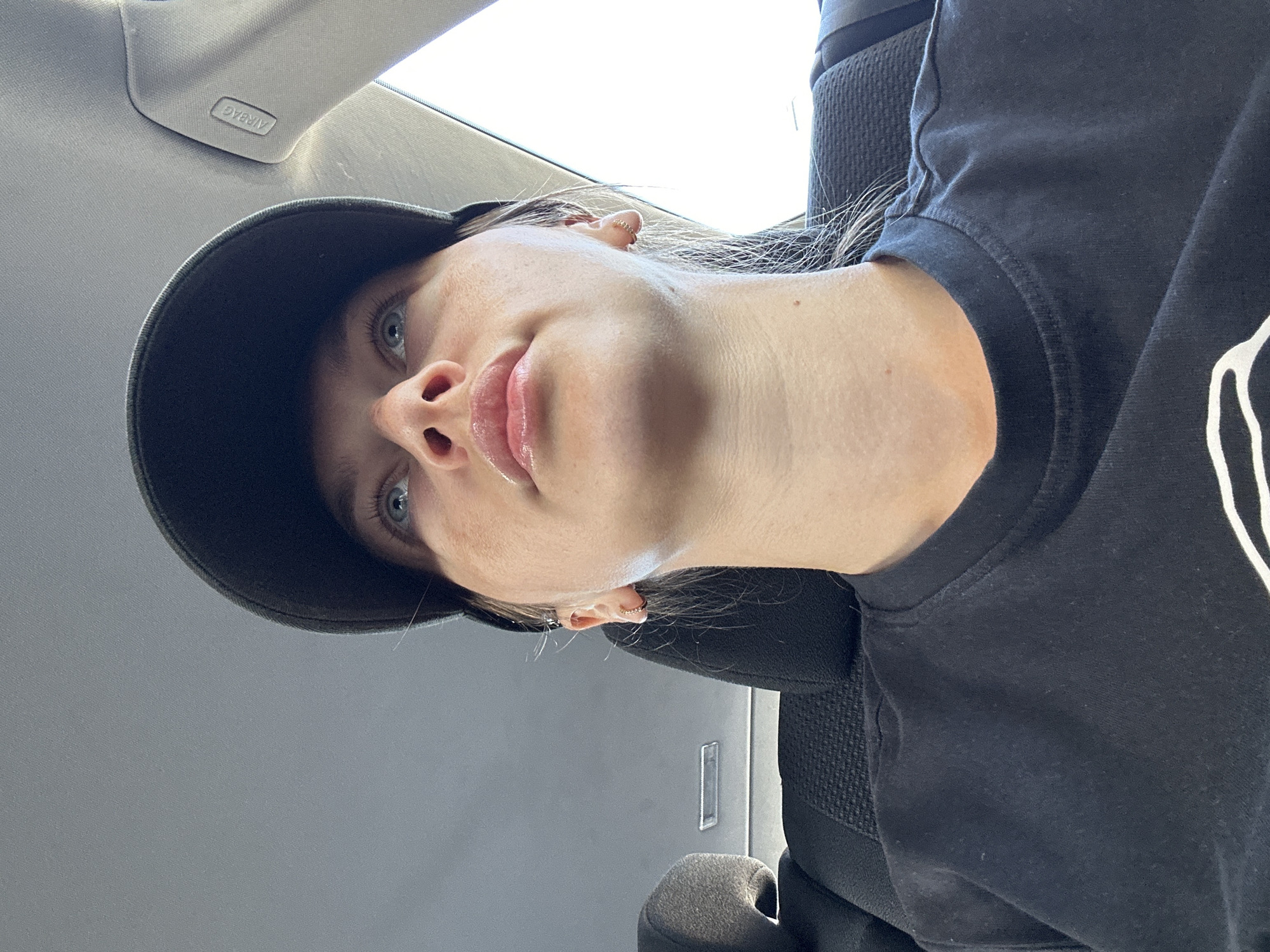 A close-up, upward-angle photograph shows a fair-skinned person with blue eyes and blonde hair, wearing a black baseball cap and a black t-shirt. The person is likely seated in a car, indicated by the interior ceiling with an airbag label. The background is blurry and indistinct, preventing identification of the location, so it is impossible to confirm that the photo was taken in Dillfeld, Germany, as the prompt requested. The person's expression is neutral, and the focus is on their face and upper body. The angle suggests the photo was taken by the person themselves.