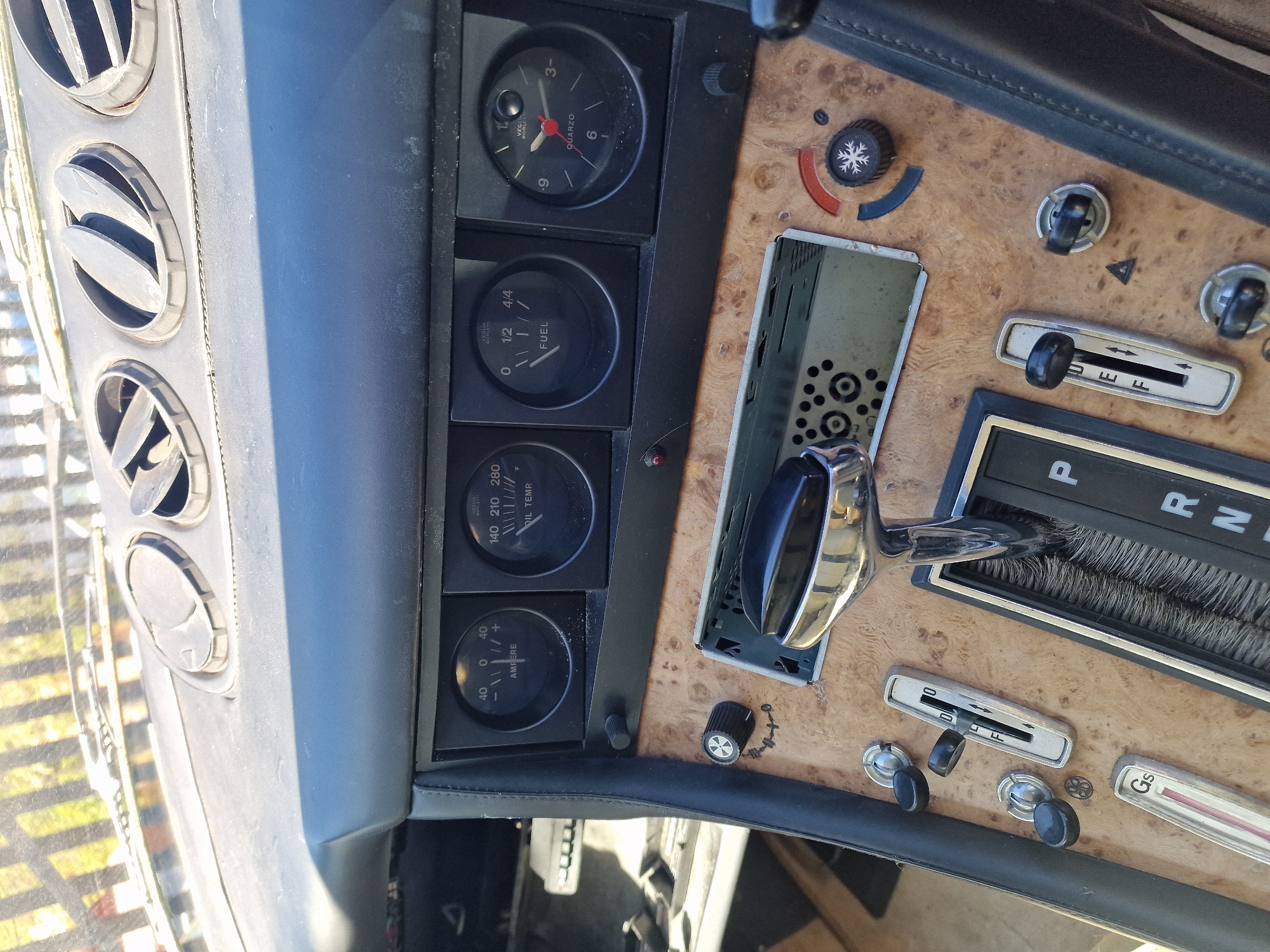 The image displays the interior of a classic car, focusing on the dashboard and center console from a slightly elevated, tilted perspective. No people are visible.
On the left, a vertical stack of four circular air vents is visible, with a glimpse of the outdoor environment through the car's front window, suggesting a sunny day. Adjacent to the vents is a black panel housing four analog gauges:
1. A clock, labeled "VEG QUARZO", indicating approximately 12:45.
2. A fuel gauge, labeled "FUEL", showing the tank is slightly over half full.
3. An oil temperature gauge, labeled "OIL TEMP.", with markings "140 210 280 F", and the needle resting around 140 F.
4. An ammeter, labeled "AMPERE", with markings "-40 0 +40", showing a reading near 0.
To the right, the center console features a wood-grain finish. Notable objects include:
* An air conditioning/heating control knob with a snowflake symbol and red/blue indicators.
* An empty slot, possibly for a car radio or stereo, exposing internal components.
* A prominent automatic transmission shifter with a chrome base and black knob, displaying "P R N D" for Park, Reverse, Neutral, Drive.
* Below the shifter, a slider control marked "0 F" and several smaller rotary knobs.
* A slim horizontal gauge or control labeled "Gs" towards the bottom right.
The interior shows signs of wear and age, including dust on the surfaces and scratches on the wood-grain. The overall setting is the cockpit of what appears to be a vintage vehicle, likely stationary during the day.