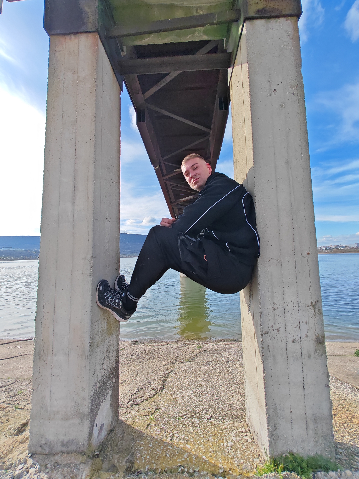 A young man sits perched on a concrete support pillar beneath a bridge. He wears a black tracksuit with white accents and black and white sneakers. The background includes a body of water, likely a lake, with land visible on the opposite shore. The sky is partly cloudy with blue patches. The location is likely the Shumen Lake Park in Shumen, Bulgaria, given the environment and construction style of the bridge.
