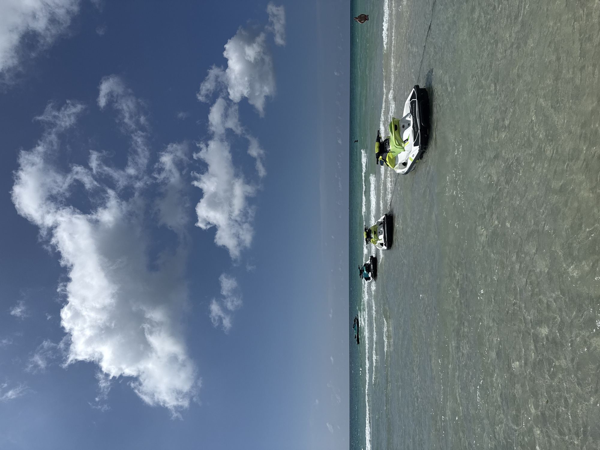 This is a vertical, medium-range shot taken from the beach looking out towards the ocean. The sky is a bright blue, with a few scattered white clouds. The ocean is a clear turquoise, with small waves breaking on the shore. In the foreground, four jet skis are parked in the shallow water. Three are on the right side of the frame, with the closest one in sharp focus. The fourth jet ski is further out in the water on the left side of the frame. A person is visible in the distance, wading in the water on the right side of the frame. The sand of the beach is not visible in this frame. The scene appears to be a typical day at a beach resort. The location is not specified in the image, but the presence of jet skis and clear turquoise water suggests a tropical or subtropical location.