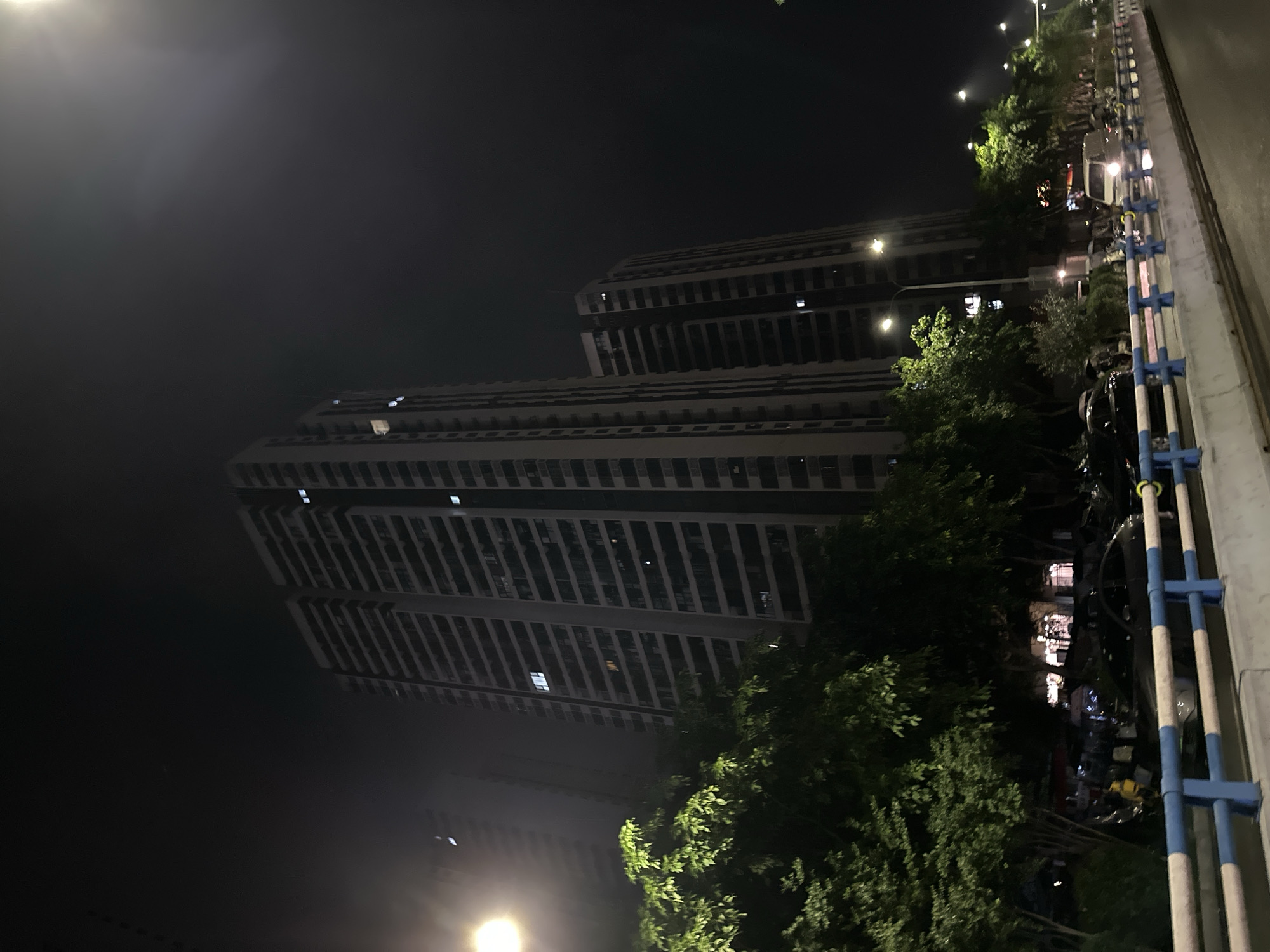 This image depicts a nighttime urban scene within the Chongqing High-tech Zone, China. The environment is dark with a high degree of atmospheric light diffusion, likely due to mist or light pollution, causing visible halos and flares around artificial light sources.
Dominating the background are multiple tall, multi-story residential apartment buildings, characterized by numerous rows of windows and balconies. The most prominent building, positioned centrally, is a light-colored structure with many illuminated windows, indicating occupancy. Several other similar buildings are visible to its left and right, receding into the darkness.
In the foreground, a multi-lane road or highway curves slightly to the right. A robust concrete barrier, painted with alternating blue and white sections and prominent blue uprights, separates the roadway from the adjacent green spaces. Lush, dense green trees line both sides of the road, partially obscuring the lower levels of the buildings and the sidewalk areas.
Artificial light sources, including streetlights and vehicle headlights, are prominent. A powerful streetlight on the far left emits a bright yellow-white glow, and other streetlights are visible further down the road on the right.
Several vehicles are present: a dark-colored sedan is partially visible on the left side, near the barrier and behind the trees, and a white van along with other indistinct vehicles are discernible on the road further down to the right. A few indistinct human figures can be observed on the left side of the scene, behind the barrier and trees, near parked vehicles. Their actions or specific features are not discernible due to the low light conditions and distance.