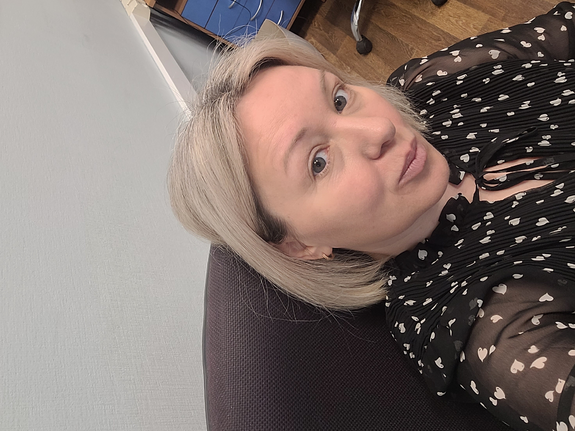 The image shows a fair-skinned woman with blonde, shoulder-length hair. She is wearing a black blouse with white heart patterns. She is seated in a dark gray chair. The background includes a light gray wall, a wooden floor, and a glimpse of a blue storage cabinet. The location of the photograph cannot be determined as Izhevsk, Russia.