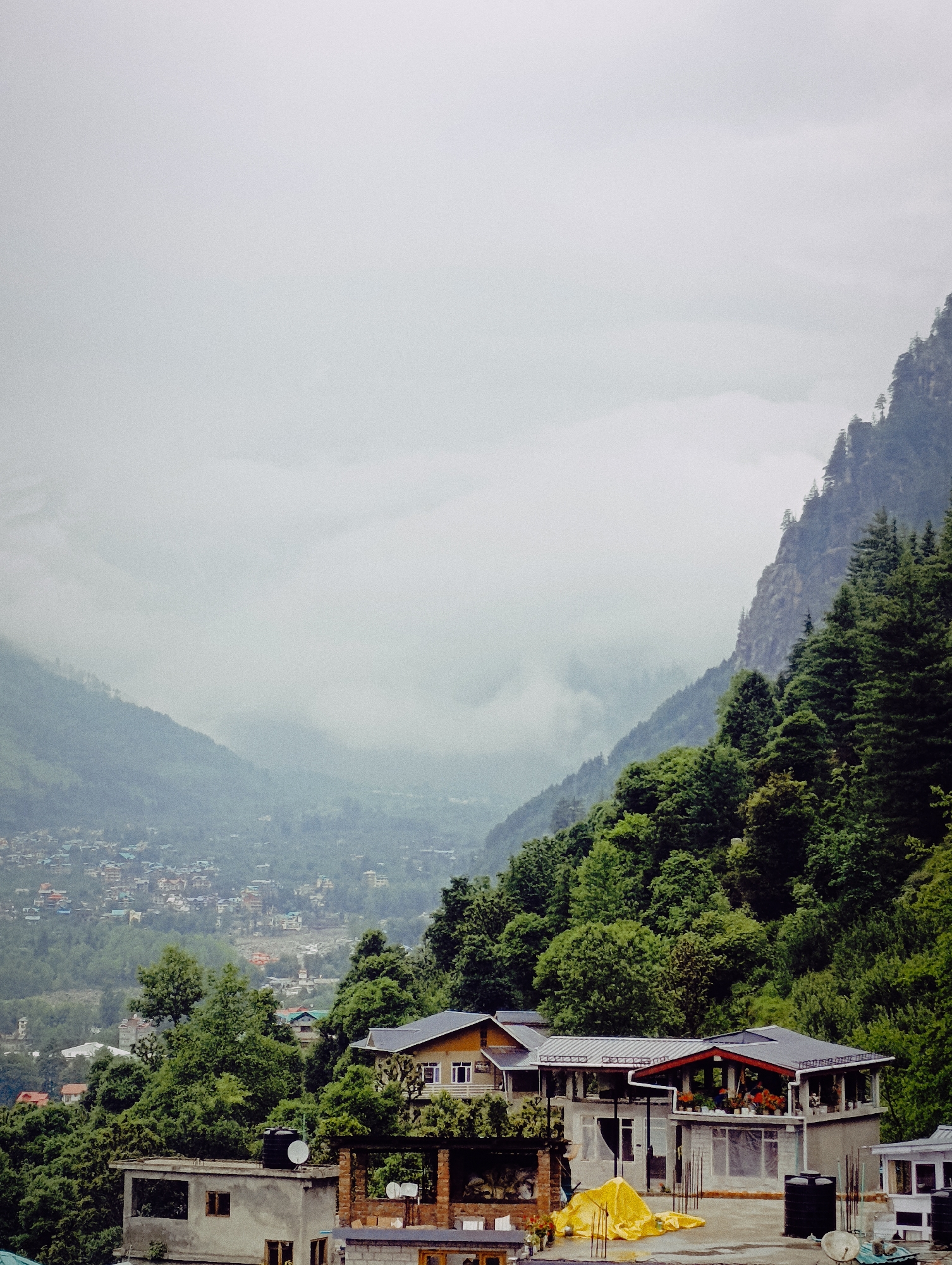 The image shows a cloudy, mountainous landscape in Vashisht, India, likely taken from an elevated vantage point. In the foreground, several multi-story buildings with flat roofs are visible. One building in the center has a dark grey roof with red trim around its upper balcony, which is adorned with potted plants. A bright yellow tarp is spread out on the concrete rooftop of a lower building to the left of this.
In the middle ground, lush green trees cover the slopes of the mountains. A distant town or village is nestled in the valley, with many small buildings visible amongst the trees. The background is dominated by towering, mist-shrouded mountains, their peaks obscured by clouds. The overall atmosphere is subdued due to the overcast weather. No people are visible in the frame.
