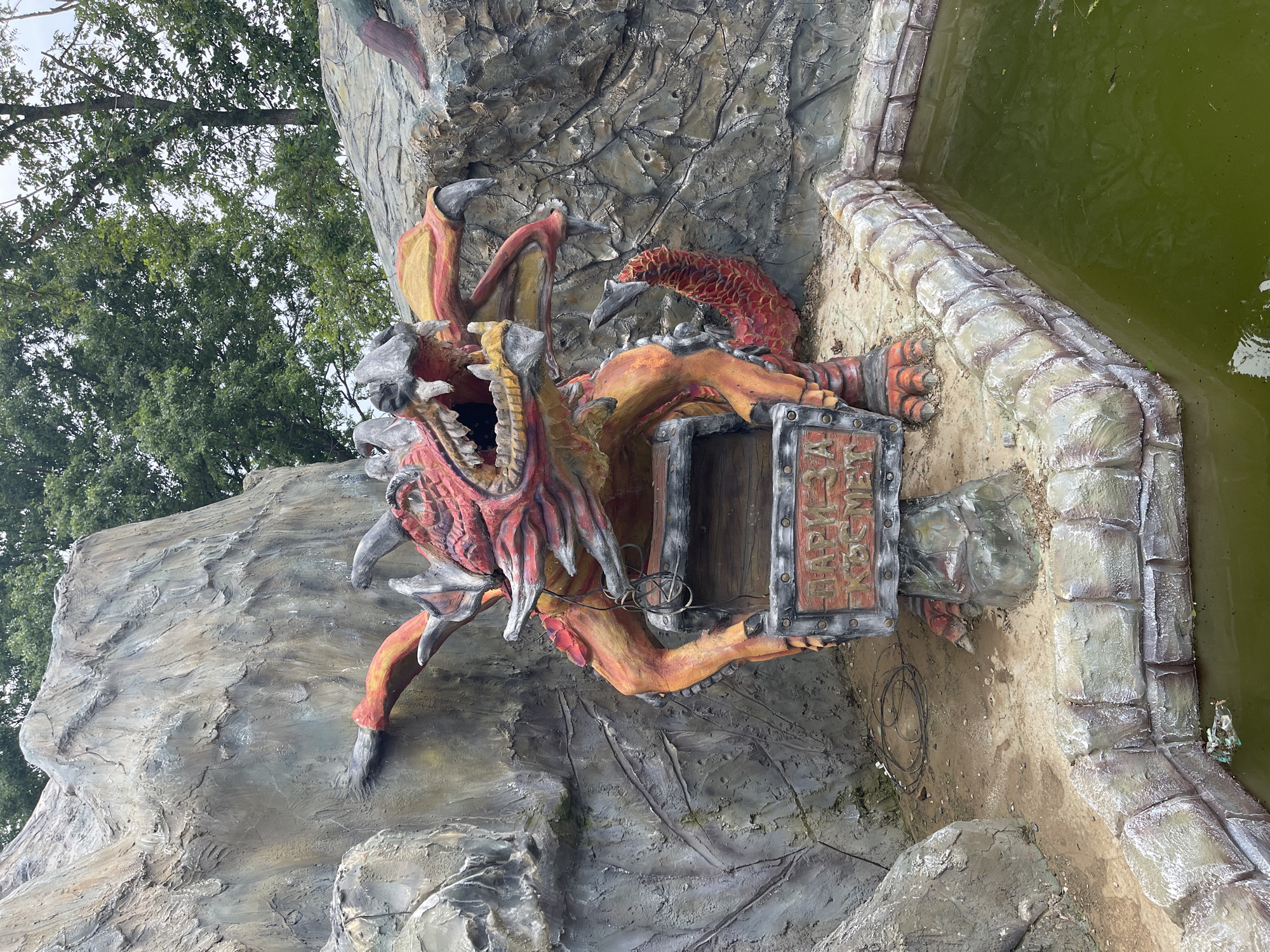 The image captures an outdoor, fantasy-themed sculptural display located in Koprinka, Bulgaria. A large, multi-colored dragon statue, predominantly red and orange with grey highlights on its claws, teeth, horns, and wing membranes, occupies the central foreground. The dragon's mouth is open, revealing fangs, and its front limbs rest upon a faux-wooden collection box. A red sign with white embossed Cyrillic lettering, reading "ПАРИ ЗА КОСМЕТ" (Pari za Kosmet), which translates to "Money for Cosmetic" or "Money for Space," is affixed to the front of the box. Black electrical wires are visible coiled on the ground near the base of the statue and the box.
The dragon is set against an artificial rock formation, textured and painted in shades of grey and brown to mimic a natural rock face. Above these formations, dense green foliage from deciduous trees is visible under an overcast sky. To the right and in the immediate foreground, a rectangular pool of stagnant, green-tinted water is present, bordered by a low wall constructed from faux-stone blocks. Visible debris, including plastic and organic matter, floats on the water's surface. The overall condition of the display suggests a degree of weathering and age.