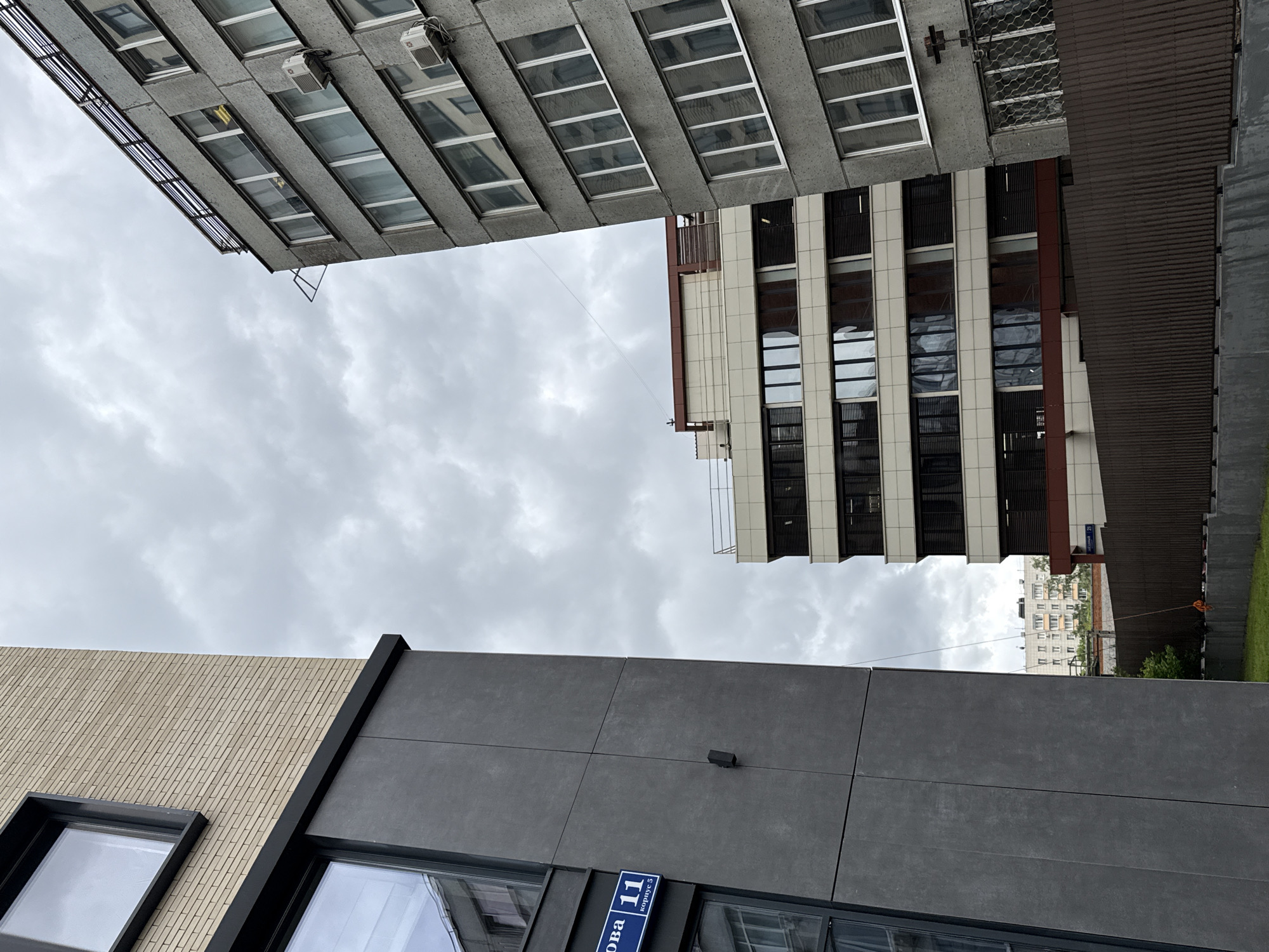 The image shows a view looking upwards at several buildings and a cloudy sky. At the top left is a gray concrete building with many windows. To the right and slightly below is a light-colored building with dark vertical stripes between its windows. At the bottom of the frame are parts of two other buildings: one with a tan brick façade and the other with a gray paneled exterior. A blue sign on the gray building reads "ул. Родионова 11 корпус 5" indicating the location as Khimki, Russia. The photograph is taken from a low angle, emphasizing the height and close proximity of the buildings. The lighting is diffused due to the overcast sky. There are no people visible in the frame. A thin wire can be seen stretching between the buildings.