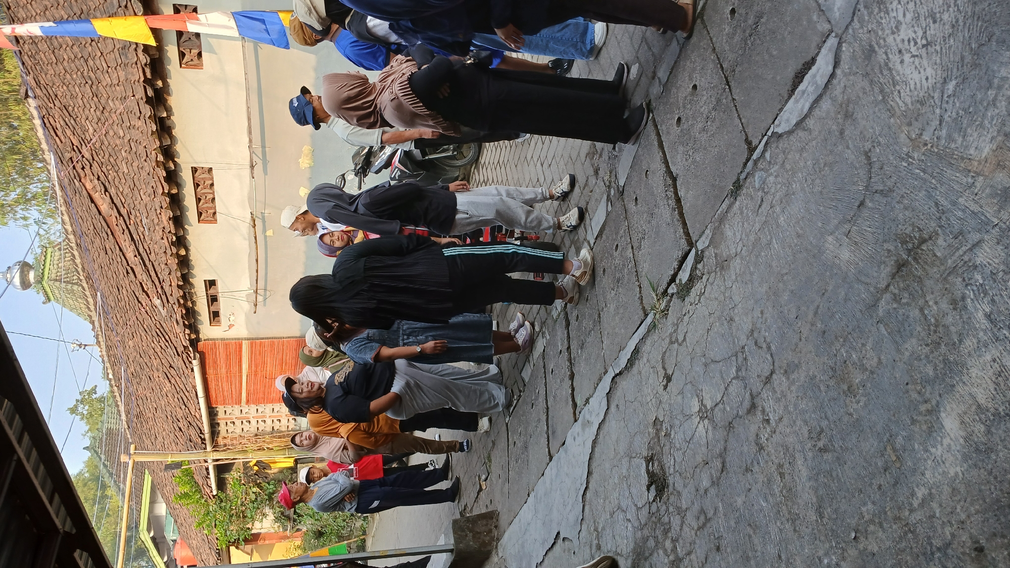 In the city of Mendut, Indonesia, a group of people stand in a line on a cracked concrete sidewalk next to a building with a tiled roof. The building appears to be a mosque, with a green dome. A flag with blue, yellow, and white colors is visible on the right side of the image. The people in the line are dressed in casual clothing, with several women wearing hijabs. Some individuals are facing forward, while others are turned to the side, suggesting they are engaged in a conversation or waiting for something. In the background, a motorcycle is parked. The sun is shining, casting shadows on the ground, indicating it is likely morning or late afternoon. The overall scene depicts a public gathering or a queue in a village setting.