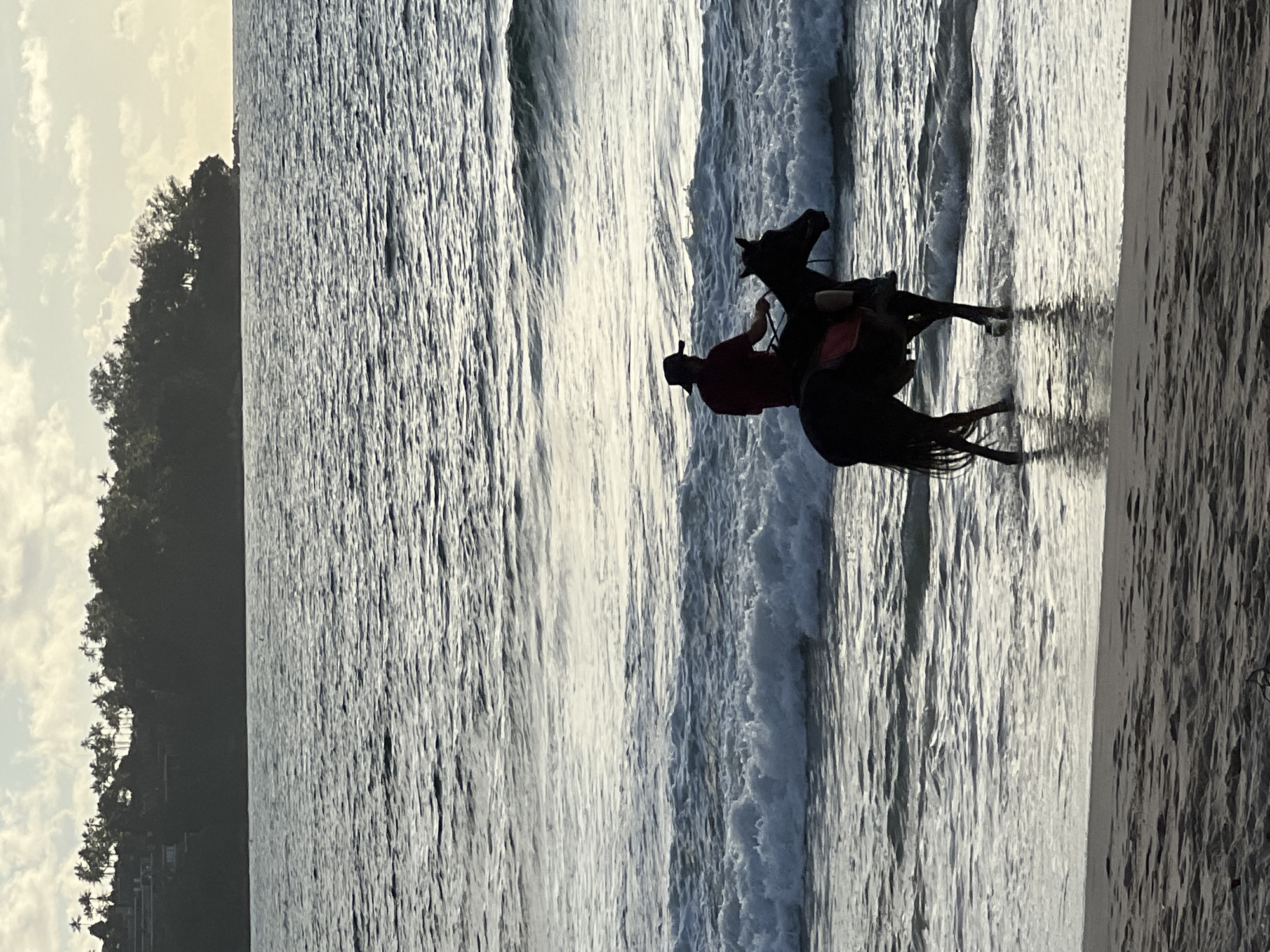 A person wearing a hat and a dark red shirt rides a dark horse along the shoreline of a beach in Choeng Thale, Thailand. The horse and rider are in shallow water with gentle waves breaking around them. The sun is low in the sky, casting a warm glow and creating reflections on the water. In the background, a tree-covered hillside with some buildings is visible under a cloudy sky. The foreground shows the sandy beach.