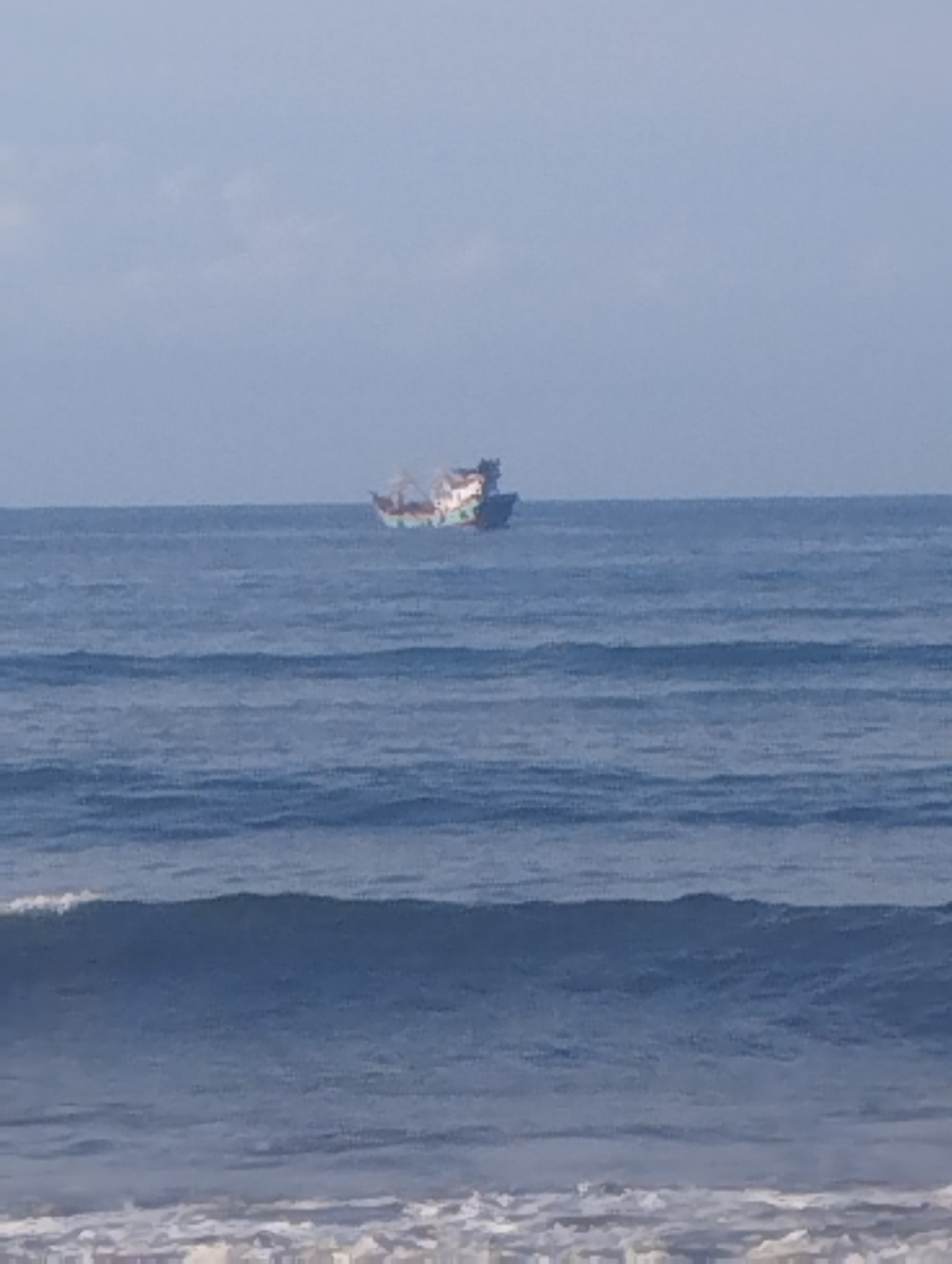 A photograph shows a ship at sea, visible from a beach. The sea is calm with small waves, and the sky is light blue with some clouds. The ship is in the distance. The location is Lhokseumawe, Indonesia.