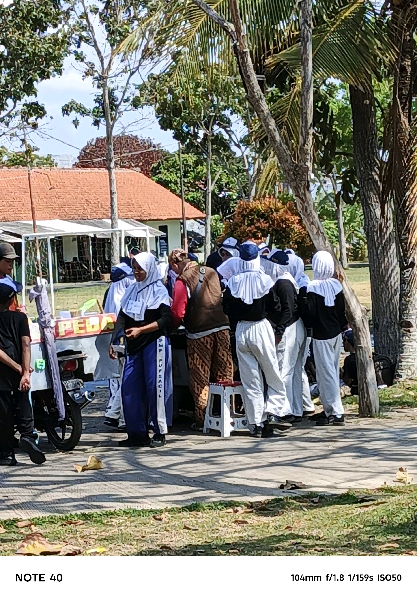 In Cilacap, Indonesia, a group of individuals, most wearing white headscarves and dark clothing, are gathered outdoors. They appear to be standing in front of a food cart, which has the word "PEBAC" visible on its side. The environment includes trees and a building with an orange roof in the background. Several people are interacting, with some looking towards the cart and others conversing. The ground is paved, with some grassy areas and fallen leaves. The lighting suggests it is daytime.