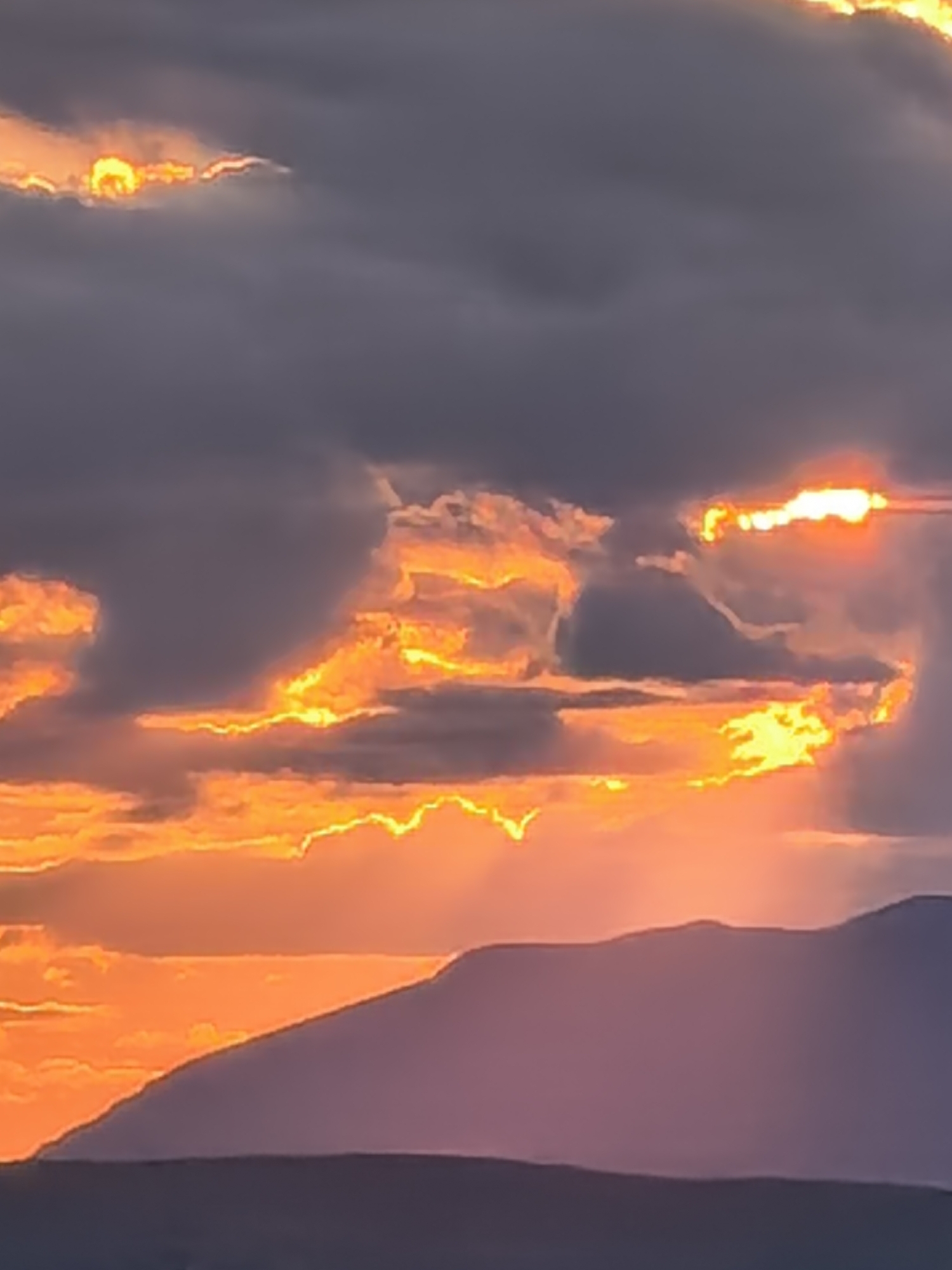 The image depicts a sunset with clouds illuminated in hues of orange and yellow. A mountain range is visible in the foreground, silhouetted against the bright sky. The location appears to be in the city of Unknown, Unknown.