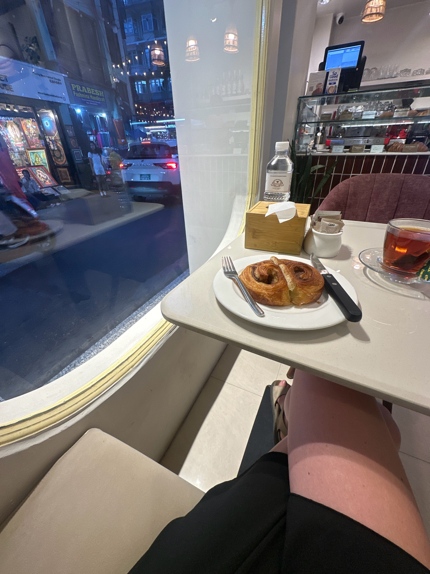 The image is an indoor scene from what appears to be a cafe in Kathmandu Metropolitan City, Nepal, with a street view visible through a large window. On the table in the foreground, a pastry, possibly a cinnamon roll, sits on a white plate next to a fork and a knife. A glass of tea, a bottle of water, and a small container of sugar packets are also on the table. In the background, through the window, a street scene unfolds with shops displaying goods, people walking, and cars on the road, illuminated by streetlights and shop lights. A white SUV with its taillights illuminated is visible. Inside the cafe, a display case with pastries and a computer monitor are partially visible. The overall lighting suggests it is evening or night.