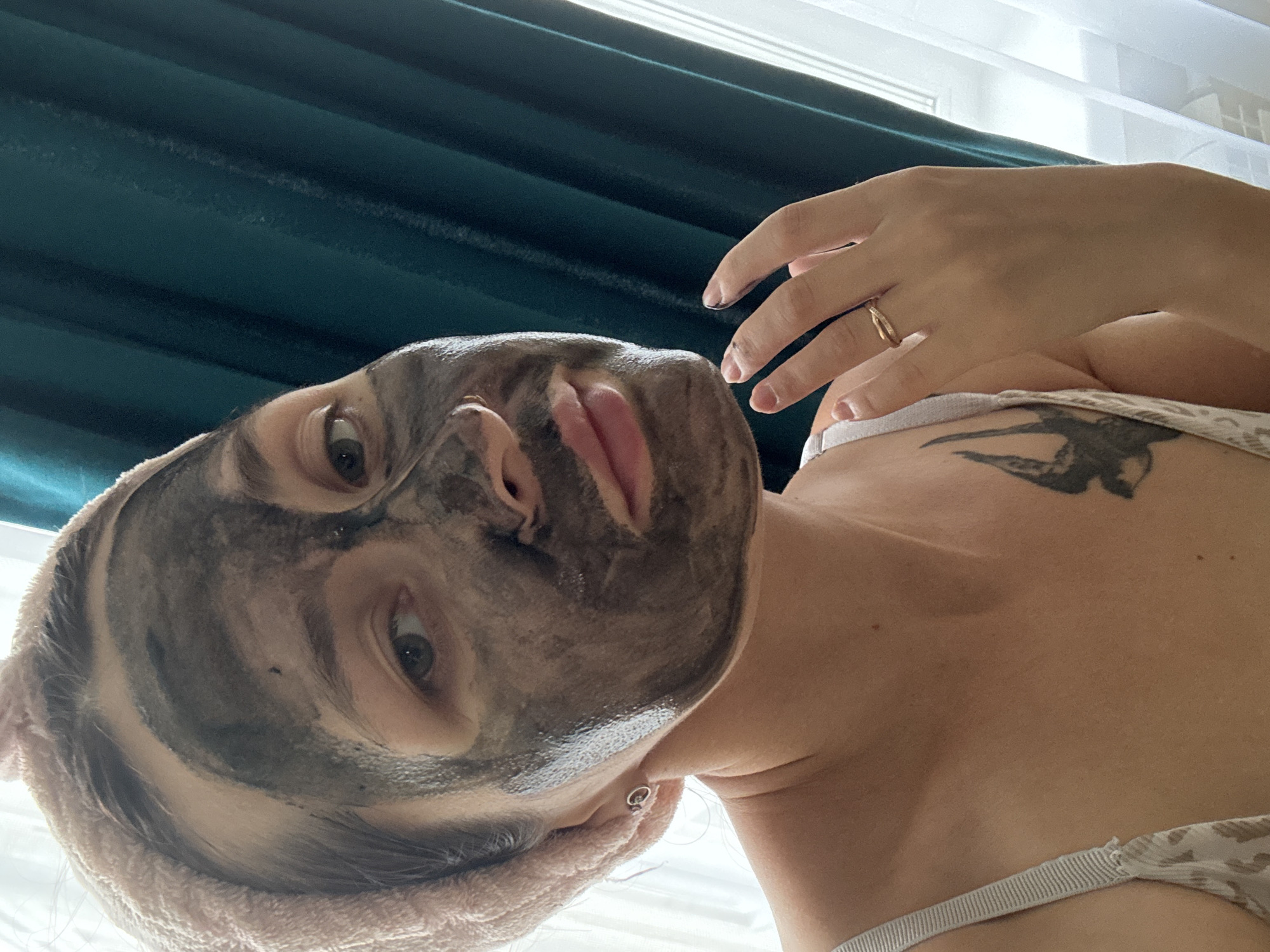 A woman with a pink towel wrapped around her head and a charcoal face mask applied to her face is posing for a selfie indoors, near a teal curtain and a window. Her left hand, adorned with a ring, is touching her face. She is wearing a light-colored bra with a patterned strap. A tattoo of a bird is visible on her upper chest. The scene appears to be a personal, domestic setting within Naberezhnye Chelny, Russia.