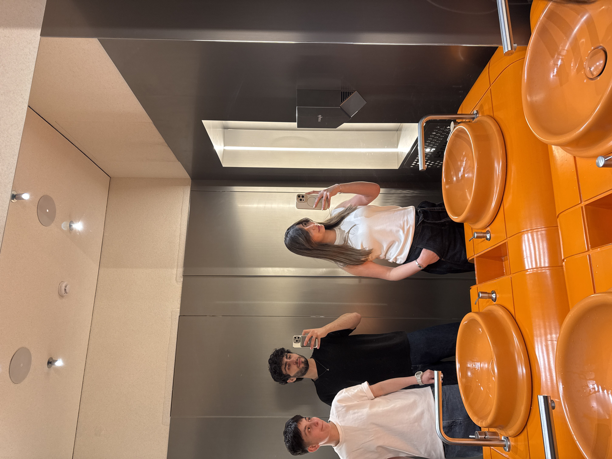 This image depicts a modern restroom or vanity area in Yerevan, Armenia, characterized by striking design elements and reflections. The photograph is oriented vertically, capturing a scene that, when viewed horizontally, shows three individuals reflected in a large mirror, along with the room's bold fixtures.
In the reflection, a woman with long, dark hair, wearing a white t-shirt and dark pants, is prominently featured, holding a light-colored smartphone up to take a selfie. To her left (in the reflection), two men are also visible. One man with dark, curly hair and a beard, wearing a black t-shirt and dark pants, is looking towards the mirror, holding a smartphone. The third person, a man with short, dark hair, is wearing a white t-shirt and dark pants, also looking towards the mirror. All three appear relaxed, engaged in taking mirror photos.
The right side of the image features a row of glossy, bright orange, modern-style sinks or urinals mounted against a dark, possibly black or dark grey, wall. Each fixture is equipped with sleek chrome faucets or flush mechanisms. Above these fixtures, a rectangular recess with integrated strip lighting illuminates the dark wall. A black dispenser, possibly for soap or hand drying, is mounted on the dark wall, with the brand name "ghd" visible on it. The wall reflected on the left side of the mirror is a lighter, speckled beige or off-white material, featuring recessed spotlights. The overall aesthetic is clean, contemporary, and functional, with the vibrant orange fixtures providing a strong focal point.