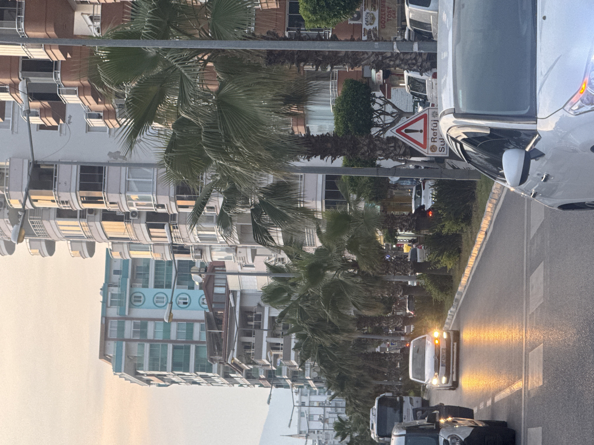 The image shows a street scene in an urban area, presumably in the city of Unknown, Unknown. The photo is taken at street level, with multiple buildings lining the left side of the street. These buildings exhibit varying architectural styles and colors, including white, beige, and turquoise. Palm trees are interspersed along the street, partially obscuring the buildings and adding a tropical touch to the setting.
The right side of the image features a roadway with visible traffic. Several cars are present, including a silver car that is positioned closely to the viewer, partially obstructing the view. Other vehicles can be seen further down the street. A white van with illuminated headlights is visible on the road.
The sky is overcast with a light, hazy color. A road sign with Turkish text and a warning symbol is situated near the curb.
