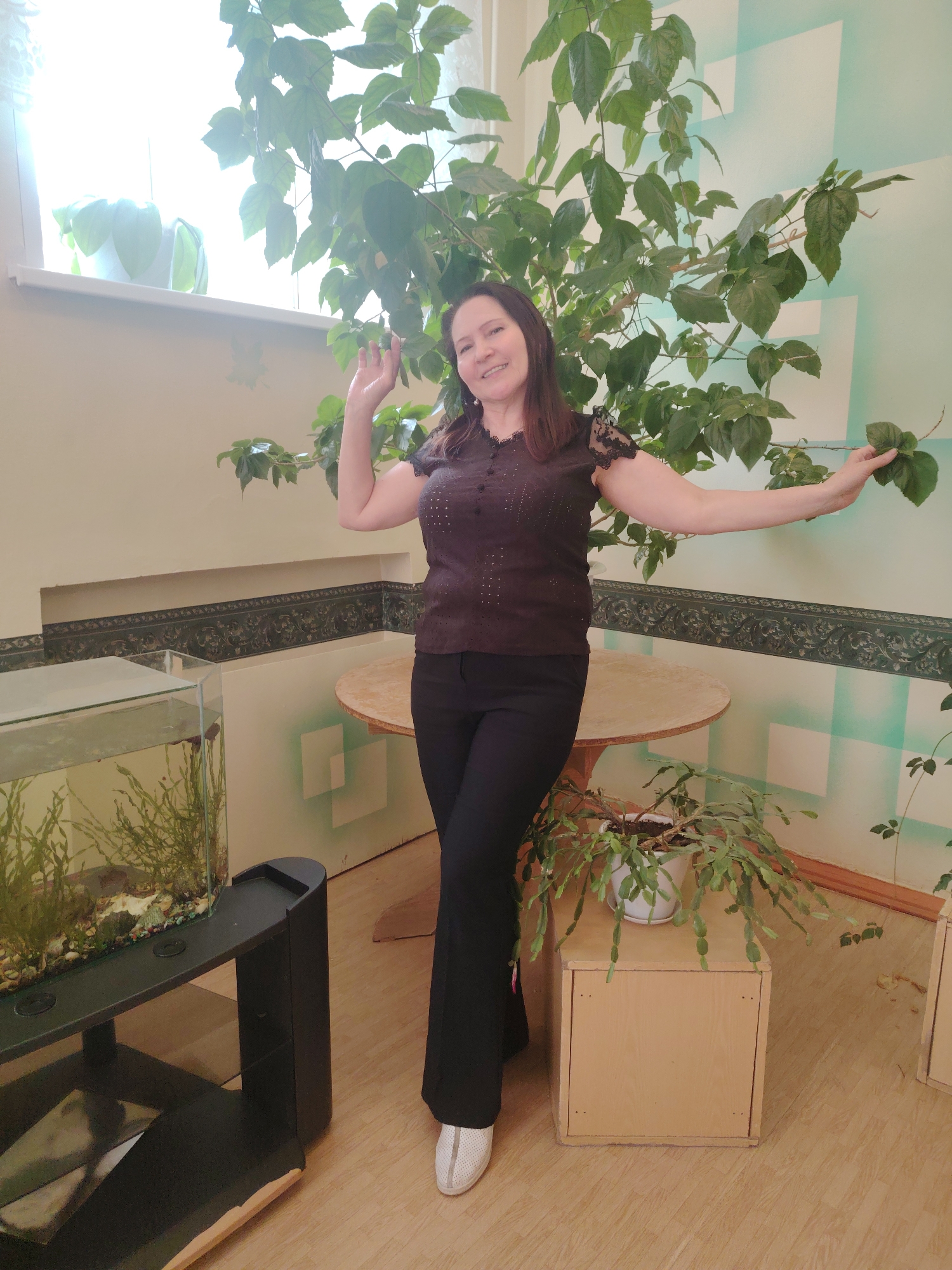 A middle-aged woman with brown hair stands indoors in Sarapul, Russia, between a large potted plant and a fish tank. She wears a dark brown, sheer blouse with lace sleeves, black pants, and white tennis shoes. Her left arm is raised, with her hand outstretched toward the plant. Her right arm is extended horizontally, holding a leaf from the plant. A round, wooden table is behind her. A fish tank filled with water and greenery is on a black stand to the left. Another potted plant sits on a small, wooden box to the right. The walls are light beige, with a decorative border. A window is visible in the upper left corner. The floor is light brown, possibly linoleum. The walls also have faint, vertical light green stripes.