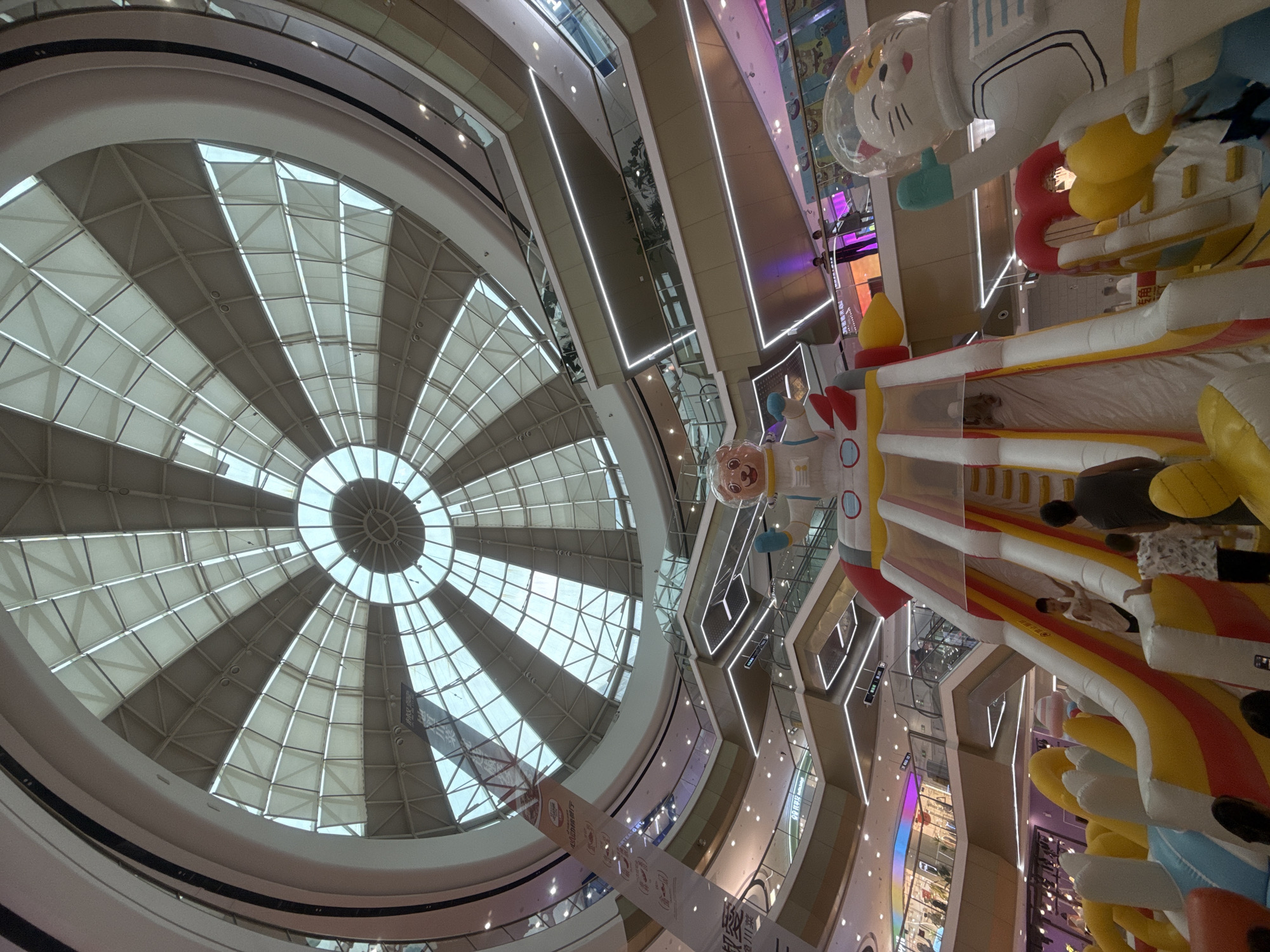 This image captures an interior view of a multi-level shopping mall, likely located in Qinhuangdao, China, given the context. The scene is dominated by a large, inflatable playground situated in the foreground. The playground features bright colors, with red, yellow, and white structures, including slides and climbing areas. One prominent inflatable is a cartoon bear in a white astronaut suit, with a red nose and big ears, positioned at the top of a slide. Another large inflatable figure is a white cat astronaut with a clear helmet.
Several people are visible interacting with the playground. Children can be seen climbing the inflatable slide and playing. Adults are also present, supervising the children. The mall interior is characterized by its modern architecture, with multiple balconies or walkways curving around the central space. These levels are illuminated by overhead lights and accent lighting strips that trace the edges of the platforms.
Above the playground and the mall levels, a large, geodesic dome ceiling made of glass panels and supporting metal structures is visible, allowing natural light to filter in. Banners hang from some of the upper levels, some featuring Chinese characters. The overall atmosphere suggests a lively and family-friendly environment within a contemporary shopping center.