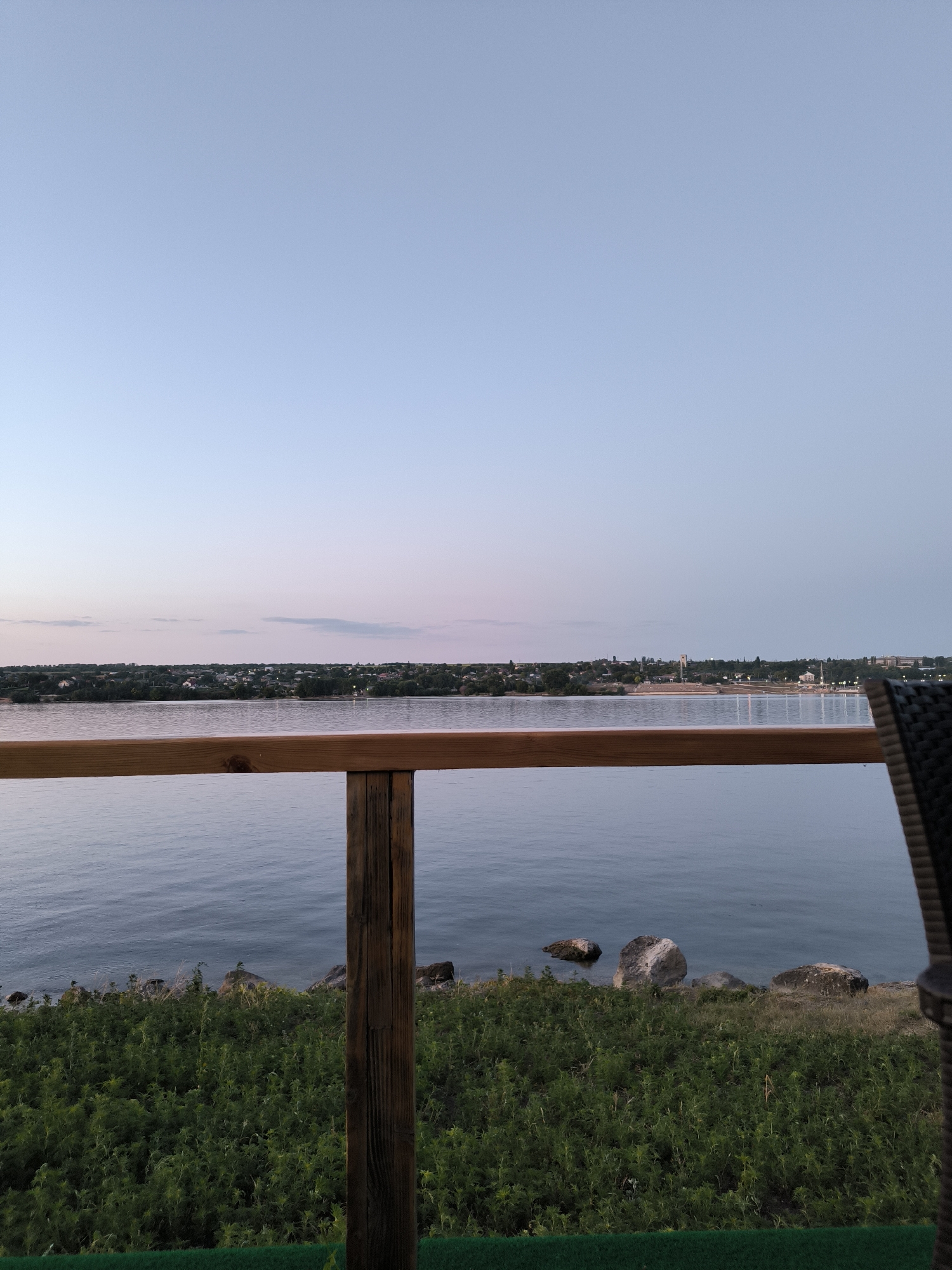 The image captures a serene waterscape at dusk, likely overlooking the Nistru River from a raised vantage point in Ustia, Moldova. The foreground features a weathered wooden railing with a vertical support post, partially obscuring a lush green embankment dotted with rocks. Beyond the embankment, a calm body of water, presumed to be the Nistru River, stretches towards the horizon. The opposite bank is lined with a dense band of trees, behind which a cluster of buildings and structures can be seen, indicative of a settlement or small town. The sky above is a gradient of soft blues and subtle pinks, typical of twilight, with a few wispy clouds. The overall atmosphere is peaceful and tranquil, with no people or discernible activity visible in the scene.