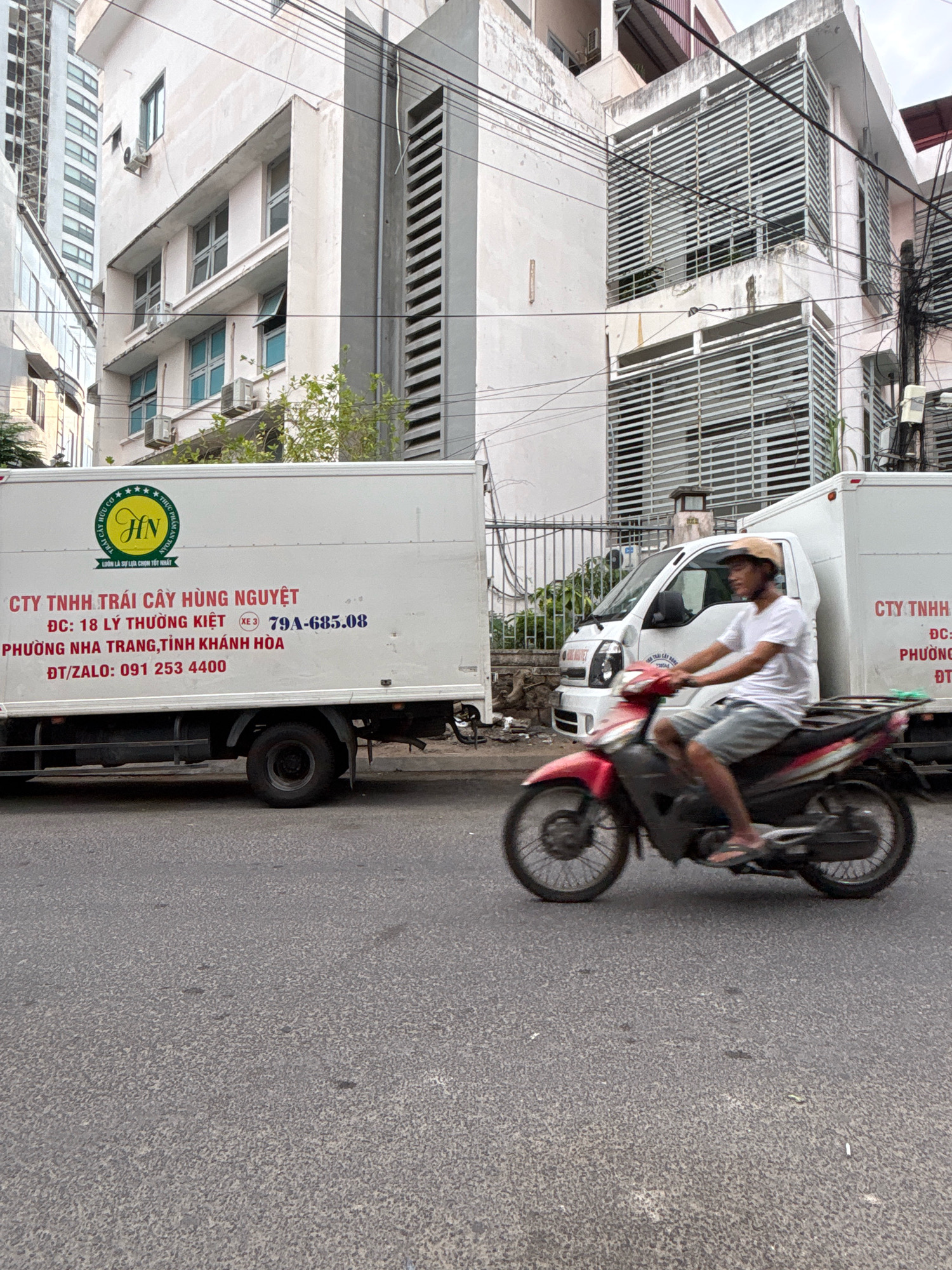 This is a medium shot, eye-level, outdoor, daytime photograph taken on a street in Nha Trang, Vietnam. In the foreground, a man wearing a light-colored t-shirt, grey shorts, and a beige hat is riding a red and black motorcycle. He is moving from left to right across the frame, with his body slightly turned towards the viewer. The motorcycle is in motion, creating a slight blur.
Behind the motorcycle, a large white delivery truck with a logo on its side is parked. The logo is a circular emblem with the letters "HN" in the center, surrounded by stars and text. Text printed on the truck in red and blue states: "CTY TNHH TRÁI CÂY HÙNG NGUYỆT", "DC: 18 LY THƯƠNG KIỆT", "79A-685.08", "PHƯƠNG NHA TRANG, TỈNH KHÁNH HÒA", and "ĐT/ZALO: 091 253 4400".
In the background, there are multi-story buildings. The building directly behind the truck is white with several windows and air conditioning units. Further back, there are taller buildings with many windows and a more modern architectural style, including one with vertical metallic blinds. Overhead, several electrical wires are visible. The street itself is paved and appears to be in moderate condition. The scene depicts a typical urban street in Nha Trang.