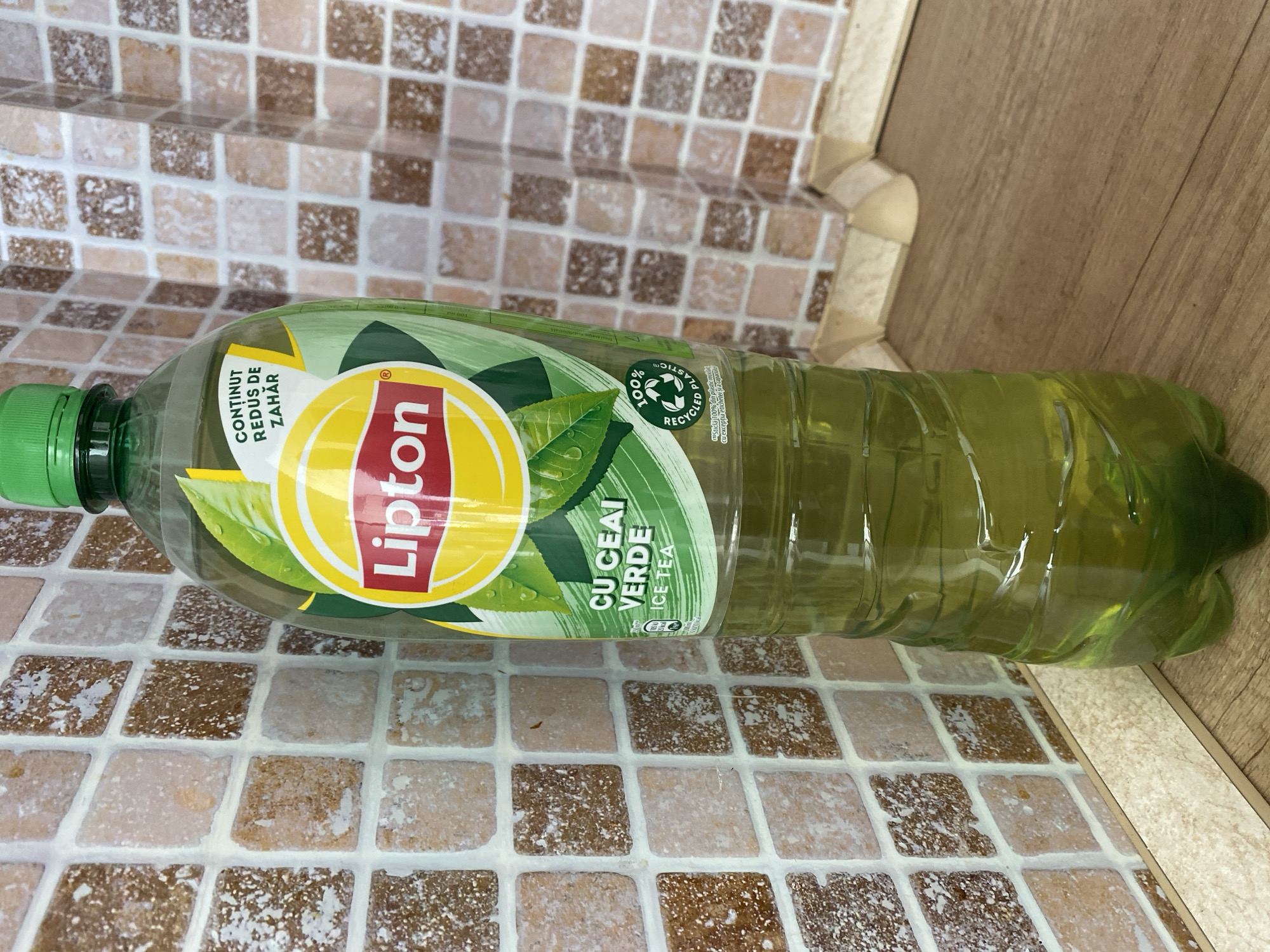 A full bottle of Lipton Ice Tea with green liquid is placed on a wooden surface in front of a tiled wall. The bottle has a green cap and a label featuring the Lipton logo, green tea leaves, and text in Romanian indicating "Cu ceai verde" (with green tea) and "Conținut redus de zahăr" (reduced sugar content). A recycling symbol with "100% recycled plastic" is also visible on the label. The tiles are a mix of brown and beige squares with white grout. The lighting suggests an indoor environment. The scene is static with no visible people or animals.