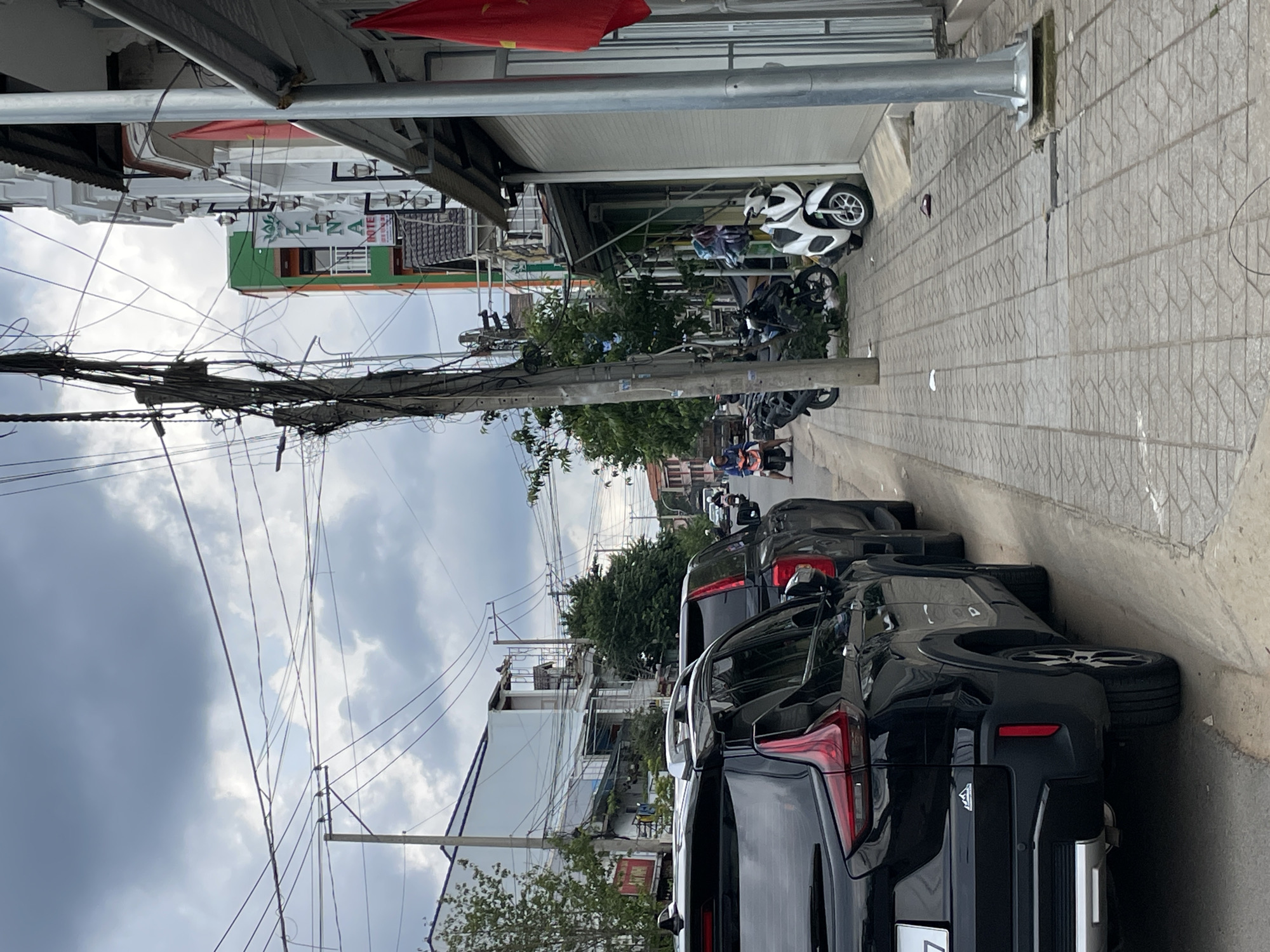 A street scene in Lâm Viên - Đà Lạt, Vietnam, features a row of parked black SUVs on the left side of the road, with a sidewalk running parallel to them. Several motorbikes are parked along the sidewalk. A light-colored motorbike is positioned in the foreground on the right. Buildings line the right side of the street, with awnings extending over the sidewalk. Electrical wires are prominently strung overhead, attached to poles. The sky is overcast with gray clouds. A red flag with a yellow star, likely the Vietnamese flag, is visible hanging from a pole on the right side of the image. A few individuals are seen on motorbikes further down the street.