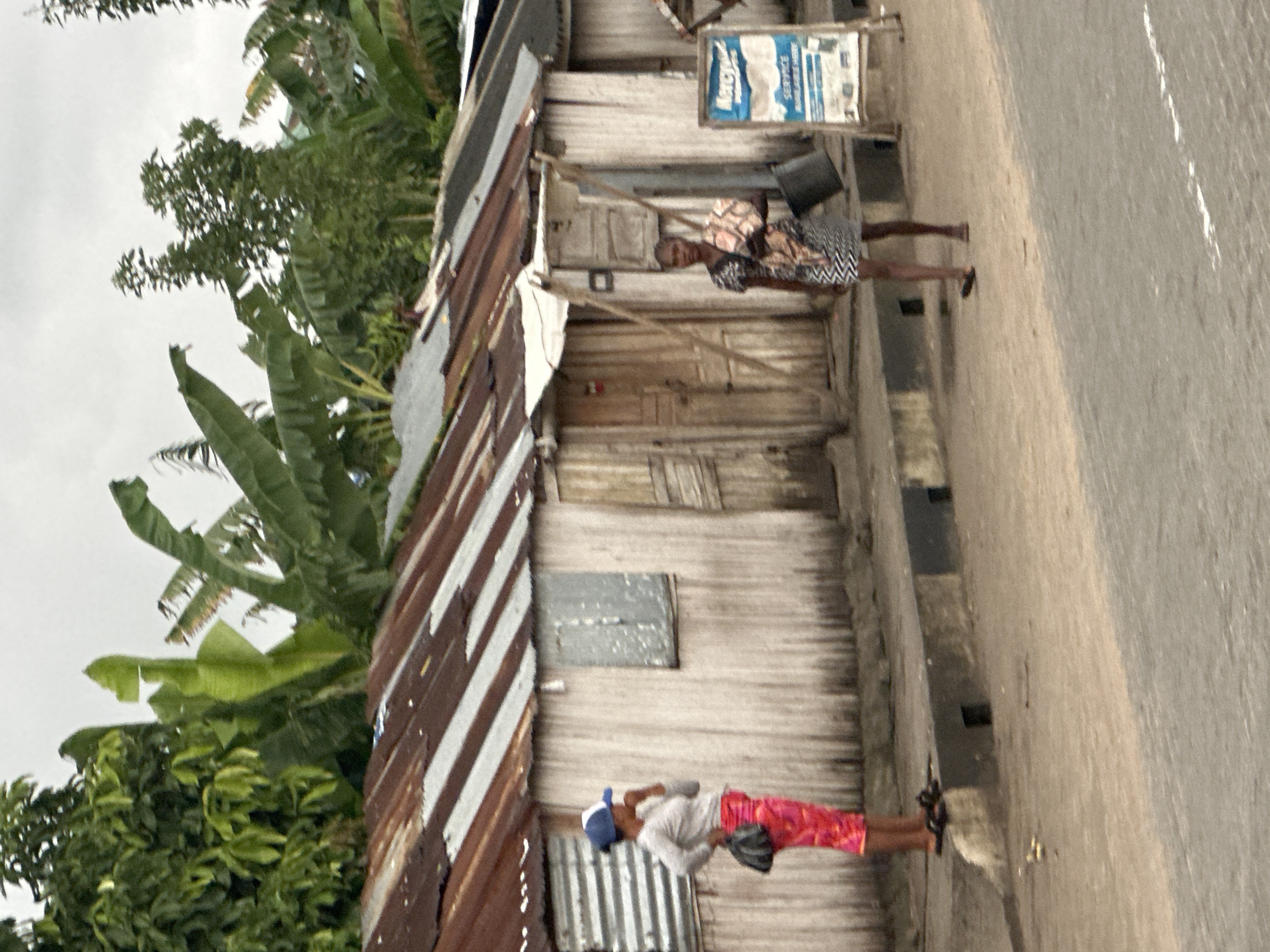 The image depicts a street scene in Badagry, Nigeria. In the foreground, a woman in a patterned dress and a zig-zag skirt walks with a bucket in her left hand and a bundle on her right shoulder. To her left, another woman stands on a curb, wearing a grey long-sleeved shirt, pink patterned pants, and a blue and white cap, with a dark handbag slung over her left shoulder. Behind them are several small buildings with corrugated metal roofs and wooden walls, some featuring simple doors and windows. Lush green trees, including banana plants, are visible in the background. A sign with blue and white lettering is positioned to the right of the buildings. The ground is a mix of dirt and paved road, with a concrete curb separating the sidewalk area from the street. The sky is overcast.