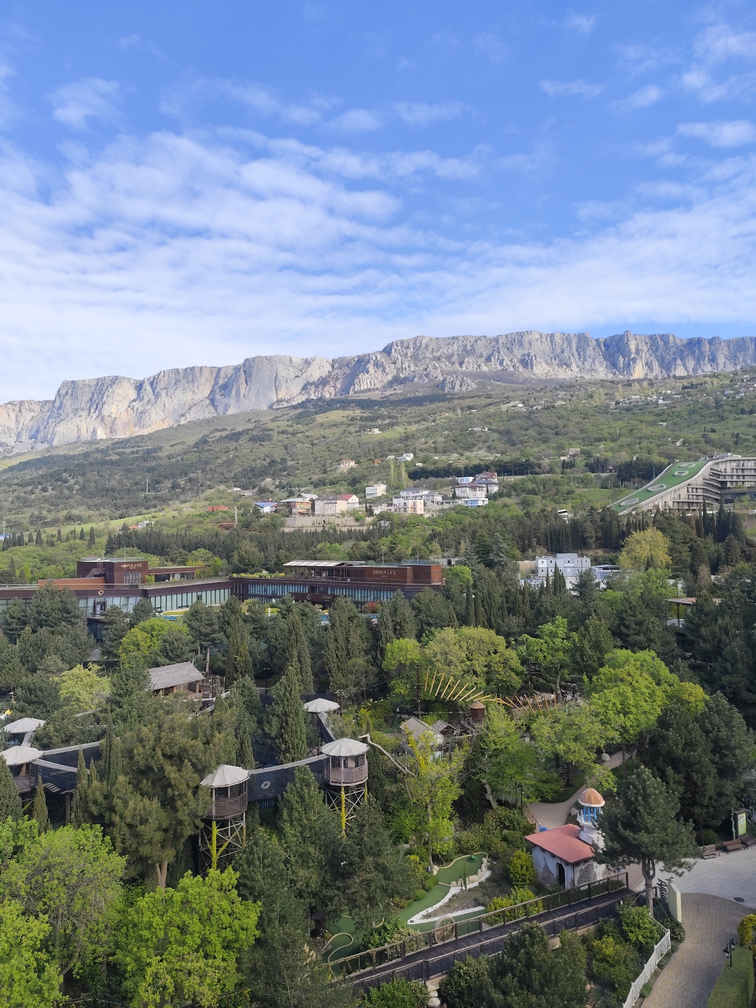 The image shows an elevated, panoramic view of Симеизский поселковый совет, Ukraine, featuring a mountainous landscape in the background beneath a partly cloudy sky. The foreground and midground consist of dense greenery, including numerous trees and shrubs. Among the trees, there is a structure with elevated walkways and towers, a minigolf course, and a building with a red roof and blue dome. Further back, a large modern building complex with multiple wings is visible, alongside several other buildings interspersed among the trees. A long, sloping structure with a green surface stretches up the hillside. The landscape transitions into a series of mountain ridges in the distance.
