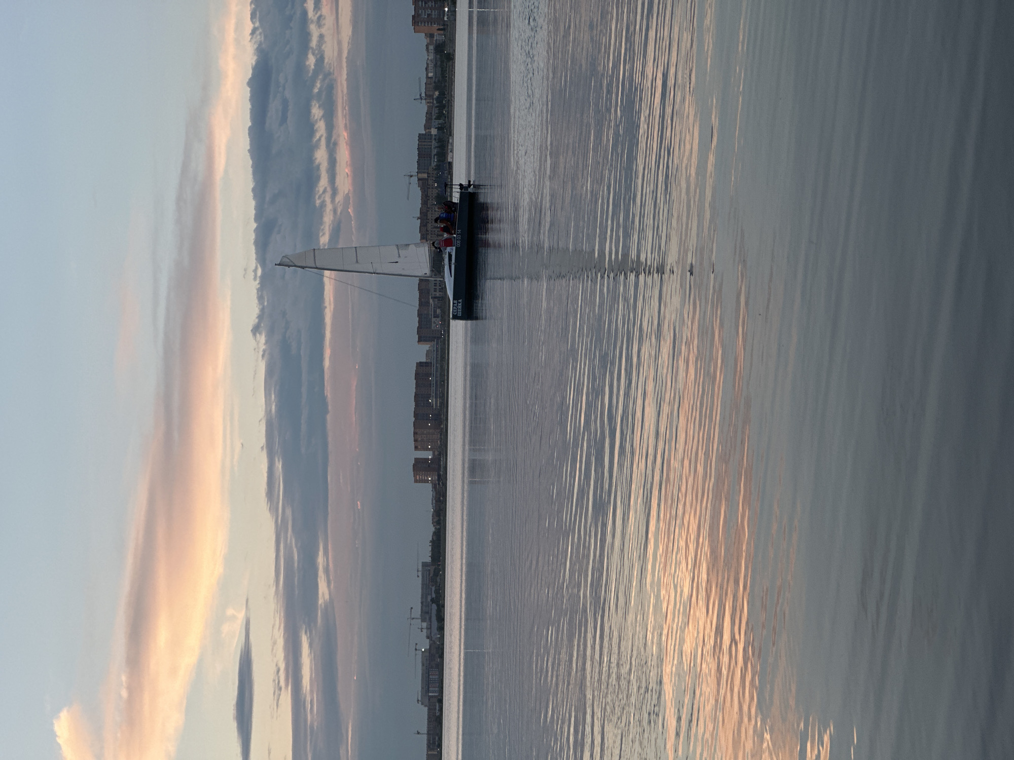 The image depicts a serene scene on a body of water, likely a lake, in Kokshetau, Kazakhstan. In the mid-ground, a white sailboat with a raised sail is navigating the water. Several individuals, possibly four or five, are visible on the deck of the sailboat. The boat has some lettering on its side, including what appears to be Russian text and numbers.
The water's surface is calm, with gentle ripples reflecting the soft colors of the sky. The sky itself is a blend of pale blue and pastel pink and orange hues, characteristic of either sunrise or sunset. Clouds are scattered across the sky, some tinged with the warm colors of the setting or rising sun.
In the background, the cityscape of Kokshetau is visible along the horizon. Buildings of varying heights, including some apartment blocks and what appear to be construction sites with cranes, form the urban skyline. The overall atmosphere is peaceful and tranquil.
