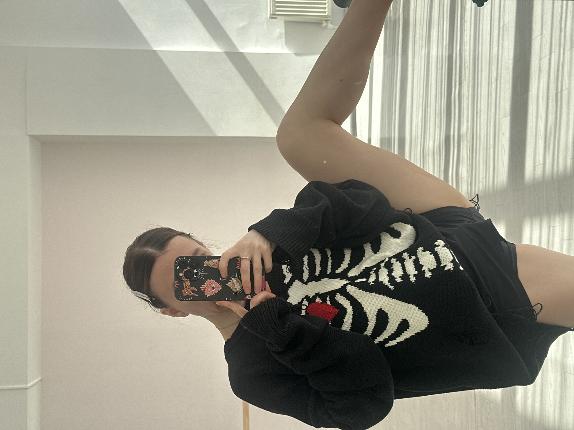 A person is captured in a mirror selfie, standing indoors against a light-colored wall. The image shows the person from approximately the head (partially obscured by the phone) down to the upper thighs.
The individual, likely female, has dark hair pulled back. She is holding a black smartphone with a distinctive black phone case featuring an array of colorful and eclectic designs, including leopard print, a pink heart, an eye, and other patterns. The phone's triple camera setup is visible. She wears a ring on one finger of her right hand, which is holding the phone.
She is dressed in a black, long-sleeved sweater that has a prominent white skeleton design on the front, depicting a ribcage and spine. A bright red heart is centered within the ribcage area. The sweater appears to be distressed or ripped in places, particularly near the hem and sleeves. Below the sweater, she is wearing black shorts. Her bare legs are visible.
The background consists of light-colored, possibly white, walls. On the right side, there is a window covered by vertical blinds or curtains, which allows bright natural light to stream in, casting distinct vertical striped shadows onto the wall and floor. In the upper background, a rectangular vent or light fixture is visible on the ceiling. The lighting suggests it is daytime. The overall scene conveys a casual, indoor setting.