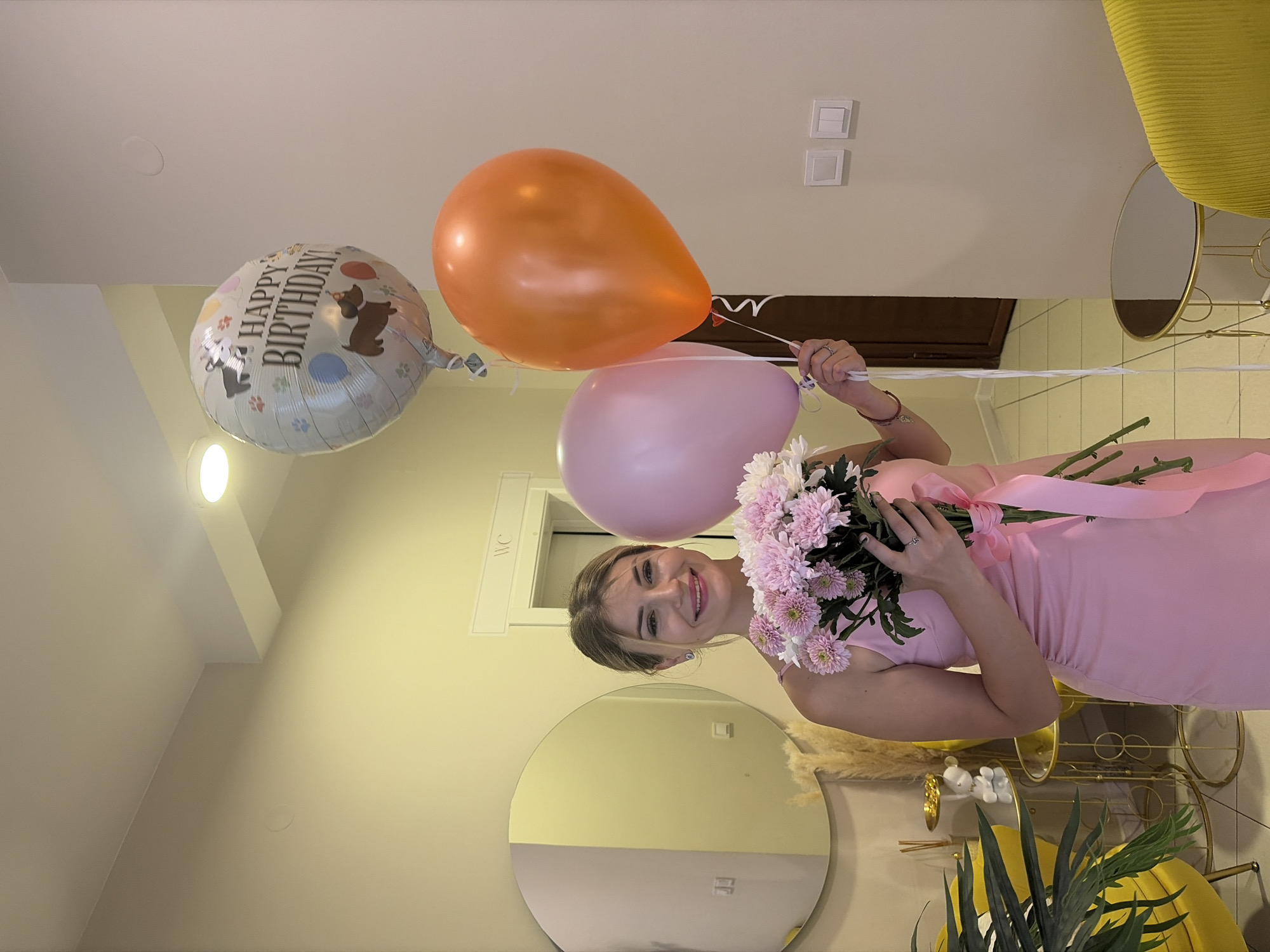 In a well-lit indoor setting, possibly an apartment or event space, a young woman with blonde hair pulled back and wearing a pink sleeveless dress stands holding a bouquet of pink and white chrysanthemums tied with a pink ribbon. She is smiling at the camera and appears to be celebrating a birthday, as indicated by three balloons she is holding. The balloons are a silver "Happy Birthday" balloon with dog illustrations, a light pink balloon, and an orange balloon.
The background features a wall painted a light beige color. To the left, a large circular mirror is mounted on the wall, reflecting a portion of the room, including a yellow armchair and some decorative dried reeds. A white sign with "WC" is visible above a doorway in the background. The floor appears to be tiled. The overall ambiance suggests a festive occasion. The provided information does not specify the location as Budva, Montenegro.