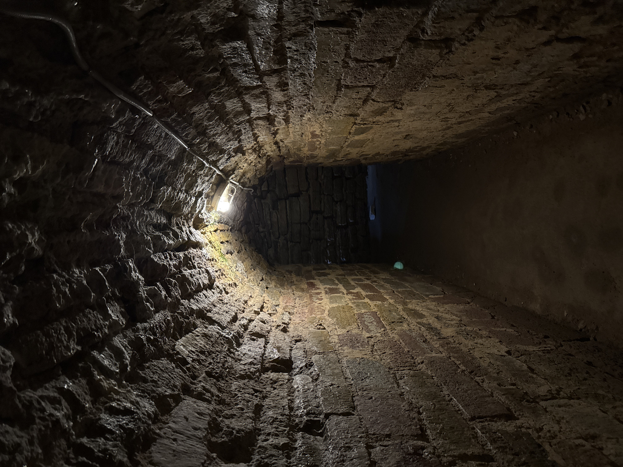This media file shows a low-angle, wide shot of a dimly lit, narrow, brick-lined tunnel. The tunnel appears to be constructed of rough-hewn stones and bricks, with evidence of dampness and possibly water on the surfaces. A single, exposed light bulb illuminates the immediate foreground, casting stark shadows and highlighting the textured, irregular surfaces of the walls and floor. A metal conduit carrying electrical wiring runs along the left wall, disappearing into the darkness ahead.
The perspective draws the viewer into the tunnel, emphasizing its confined nature and the rough, aged construction. The lighting creates a sense of mystery and perhaps slight unease, as the tunnel recedes into deep shadow. The floor is paved with bricks, also appearing wet and worn. The overall impression is one of a historic or underground passage, possibly a cellar, service tunnel, or part of an older infrastructure. There is no indication of people or specific activity, suggesting a static, architectural capture. The setting is implied to be Posyolok Imeni Morozova, Russia, but this context is not visually evident within the image itself. There is no visible text.