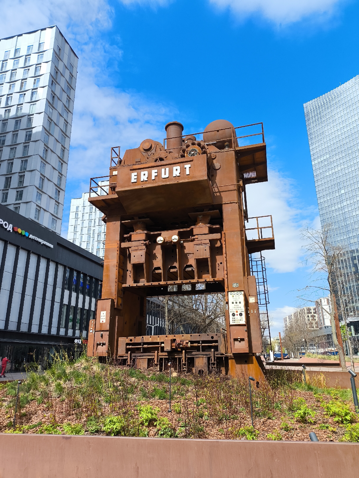 The image shows an Erfurt drop hammer monument in Moscow, Russia, set against a backdrop of modern buildings and a bright blue sky with scattered clouds. The drop hammer, a large, rust-colored industrial machine, dominates the foreground and is surrounded by a raised garden bed with various plants and low-level lighting. The machine has the word "ERFURT" in large, white letters on its front. To the left of the monument, there's a building with a black and white striped facade and the word "ГОРОД" (City) visible. A tall, white building rises behind it. To the right, another modern glass-faced building is present. The overall scene is urban, with elements of industrial heritage integrated into a contemporary environment.