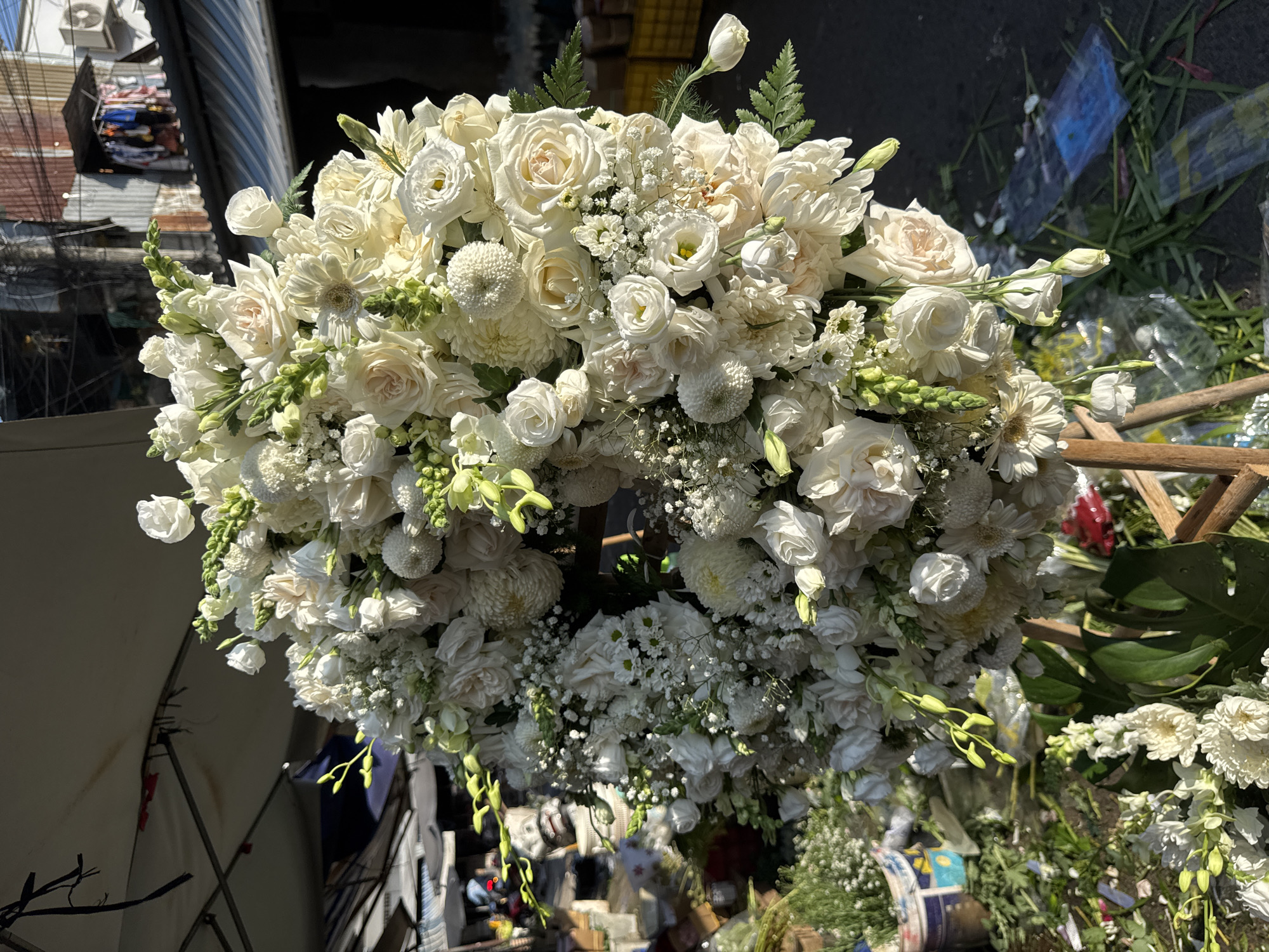 A large, elaborate arrangement of white flowers, primarily roses and chrysanthemums, is displayed outdoors on a wooden stand in what appears to be a street market or a shop entrance in Nha Trang, Vietnam. The floral arrangement is circular and densely packed, featuring various textures and sizes of white blooms, accented by small green buds and foliage. In the background, a corrugated metal roof with laundry hanging from it, an air conditioning unit, and electrical wires suggest an urban environment. The ground is paved and appears slightly wet in places, with scattered green leaves and some plastic-wrapped items. The lighting indicates it is daytime, with strong sunlight casting shadows.