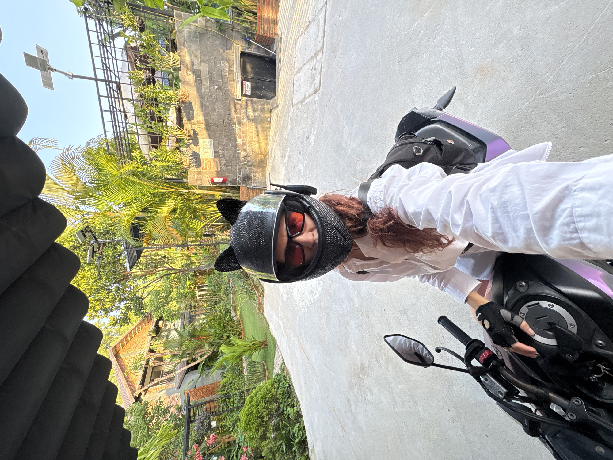 A person wearing a black helmet with cat ears and sunglasses is sitting on a motorcycle. The person is wearing a white collared shirt and black fingerless gloves. The motorcycle is black with purple accents. The background appears to be a courtyard with a brick building, lush greenery, and a clear blue sky. The person is holding the motorcycle's handlebars with their left hand and taking a selfie with their right hand. The scene is outdoors in what appears to be a tropical or subtropical environment, possibly at a resort or a private residence.