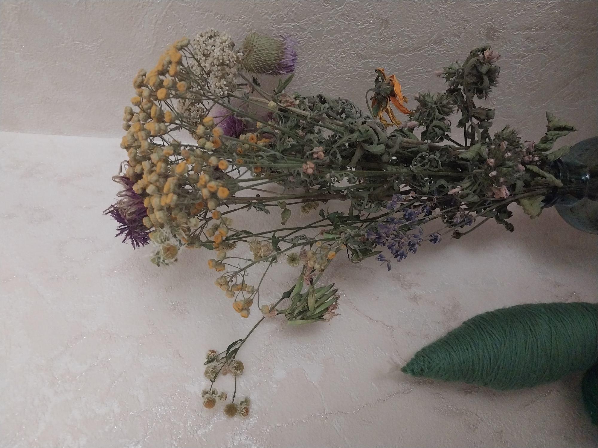 A bouquet of dried flowers is displayed in a vase against a textured, light-colored wall. The bouquet contains various dried floral elements, including small yellow flowers, purple thistles, and wispy white clusters. To the left of the bouquet, a conical object wrapped in green yarn stands vertically. The scene appears to be indoors, with the lighting suggesting an ambient light source rather than direct sunlight. No people are visible in the image.