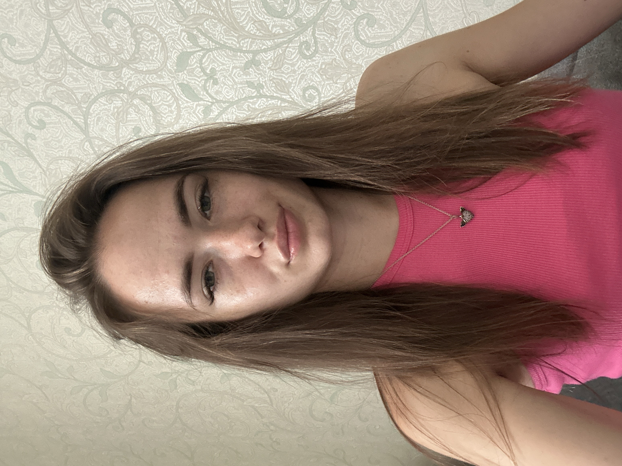 The media file is a selfie of a young woman taken indoors. She has long, brown hair and is wearing a pink tank top with a silver necklace featuring a small, heart-shaped pendant. Her skin is fair with some freckles visible. She has light-colored eyes and her lips are slightly parted. The background is a wall with a subtle, light-colored patterned wallpaper. The lighting suggests it is daytime, but no specific time can be determined. The woman's expression appears neutral or slightly amused. There is no visible text in the image.