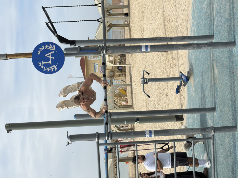 This image depicts an outdoor calisthenics gym situated on a public beach in Tel-Aviv, Israel.
In the foreground, a shirtless, light-skinned male with visible arm and chest tattoos and blonde/light brown hair is performing an advanced calisthenics maneuver, suspending his body horizontally above a set of gray metal parallel bars with his legs bent and raised upwards. He wears loose-fitting light-colored pants and a thin chain necklace.
To the left, a male with dark hair and a beard, wearing a white t-shirt, dark shorts, white socks, and white slides, stands observing. He holds the leash of a small, light-colored dog that stands beside him. Partially visible next to him is a female with a cap.
The gym features multiple gray metal poles and bars, with a blue circular sign displaying "LA" within a white laurel wreath mounted on a wooden pole. The ground beneath the gym equipment is a light blue, textured surface.
The background consists of light-colored sand extending to the sea. A multi-story lifeguard station with a red roof, white walls, and blue railings is prominently visible on the beach. Other background elements include wooden beach shelters with awnings, stacked white plastic chairs, and a yellow paddleboard. The sky is partly cloudy.