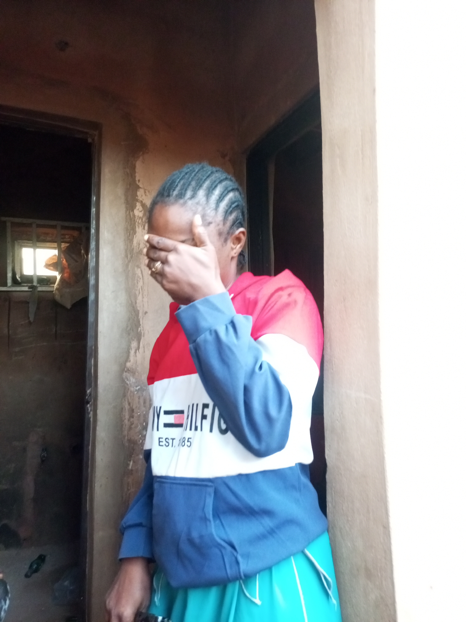 A woman stands in a doorway, covering her eyes with her right hand. She wears a hooded sweatshirt with red, white, and blue color blocking, and a bright teal skirt. The background shows a rustic, unfinished structure with a narrow window and a partially visible interior. The scene appears to be in Tempure, Nigeria.